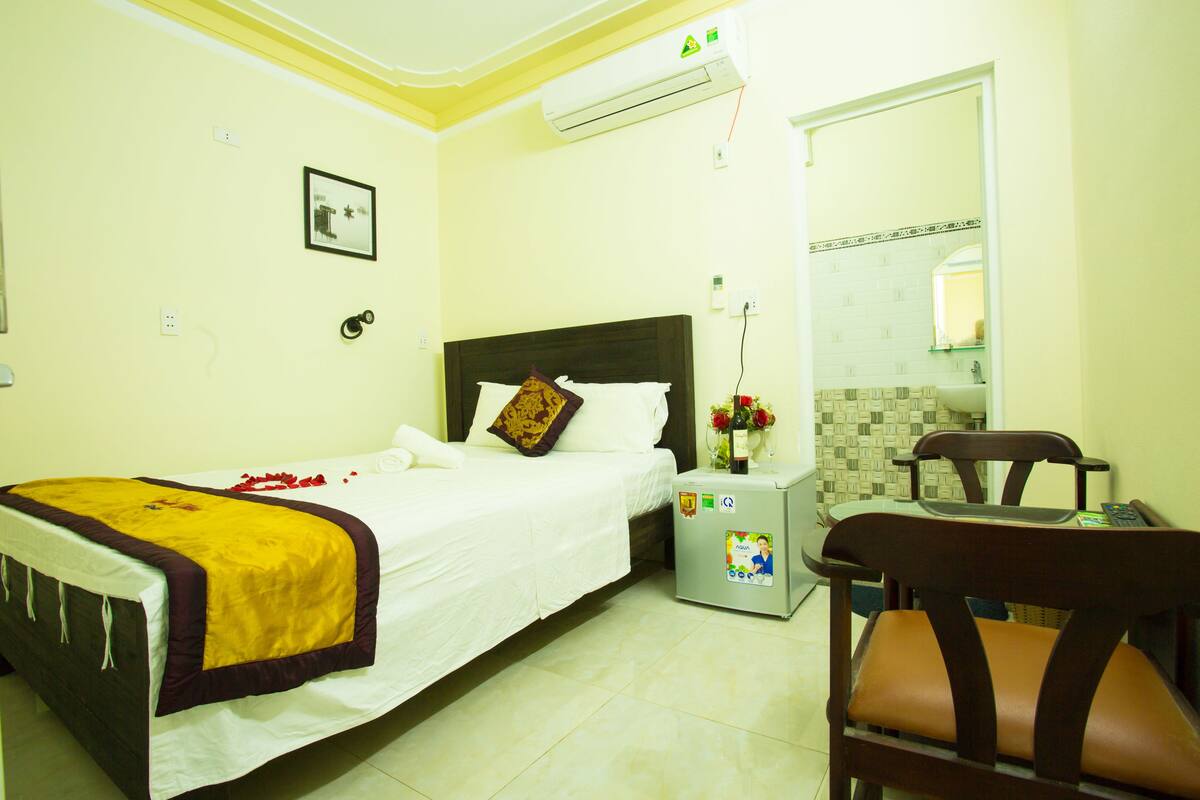 A hotel room with a bed, a nightstand with a lamp, a chair, and a bathroom visible through an open door.