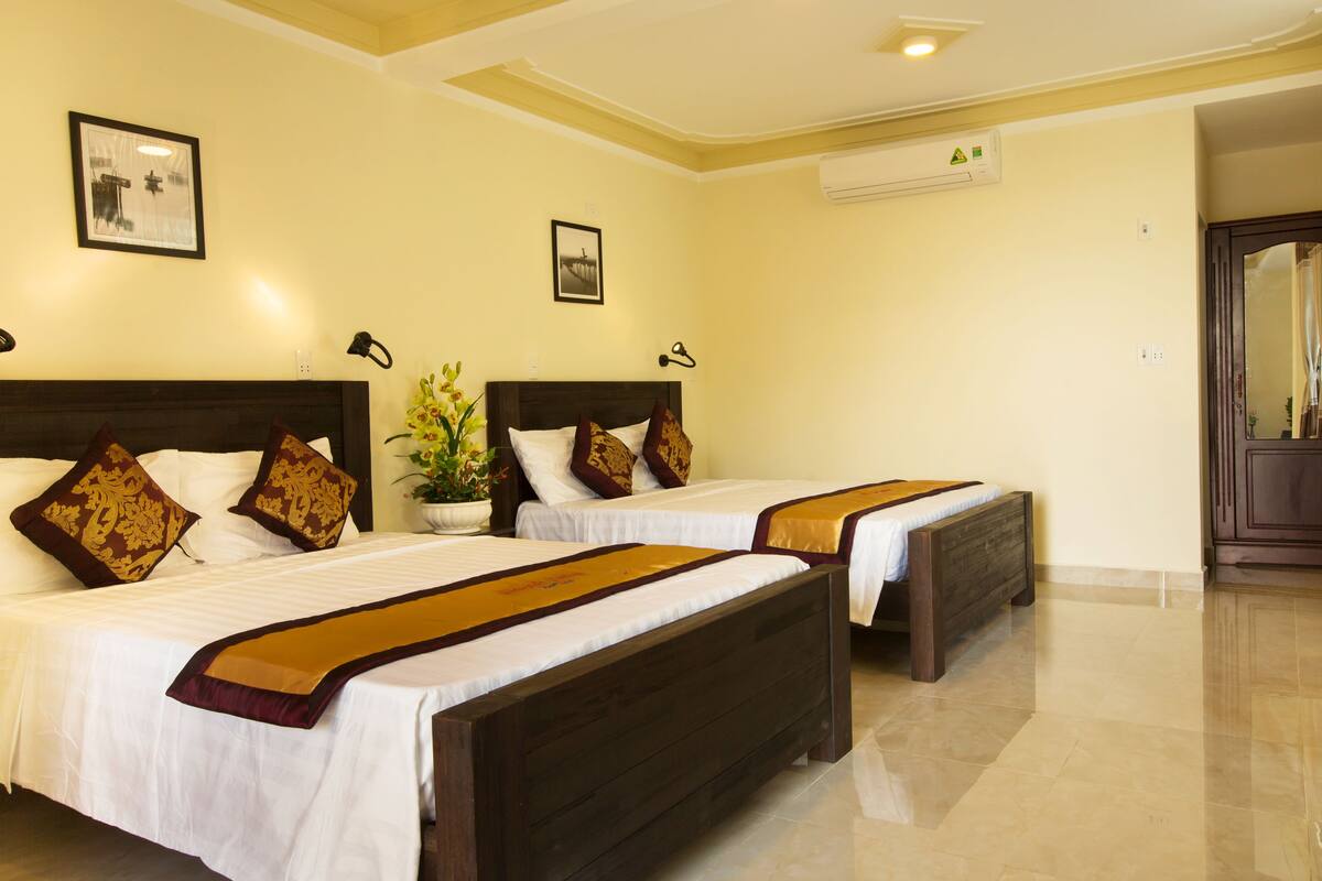 A hotel room with two beds, each with a wooden headboard, white bedding, and decorative pillows. There is a potted plant on the left side of the room, and an air conditioning unit mounted on the wall.