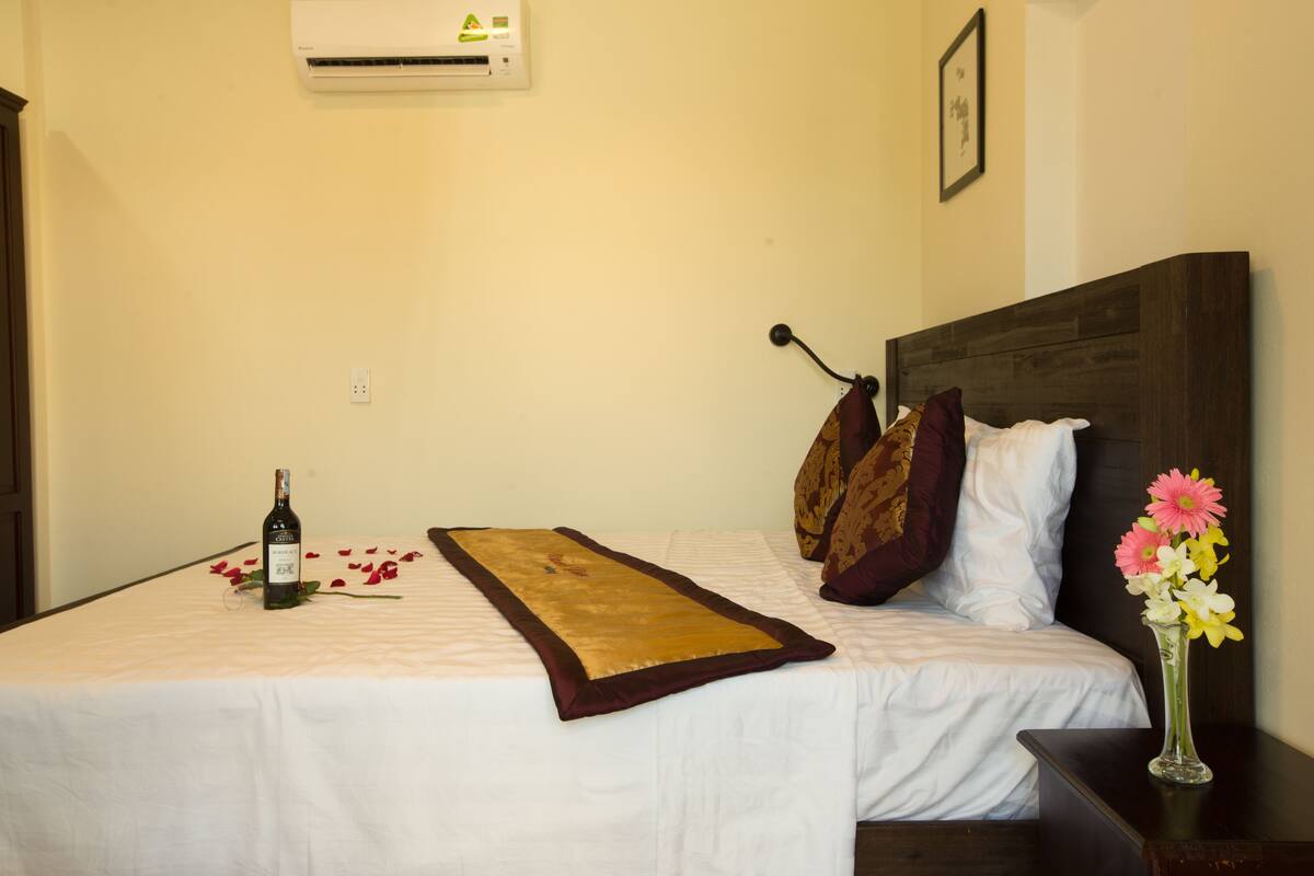 A hotel room with a bed, a bedside table, a vase with flowers, and an air conditioning unit on the wall.