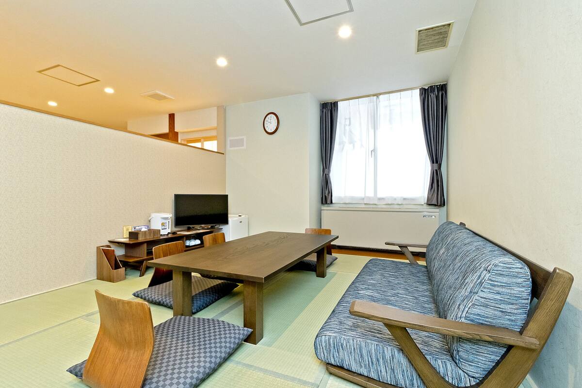 A room with tatami flooring, a wooden table, a sofa, a television, and a clock on the wall.