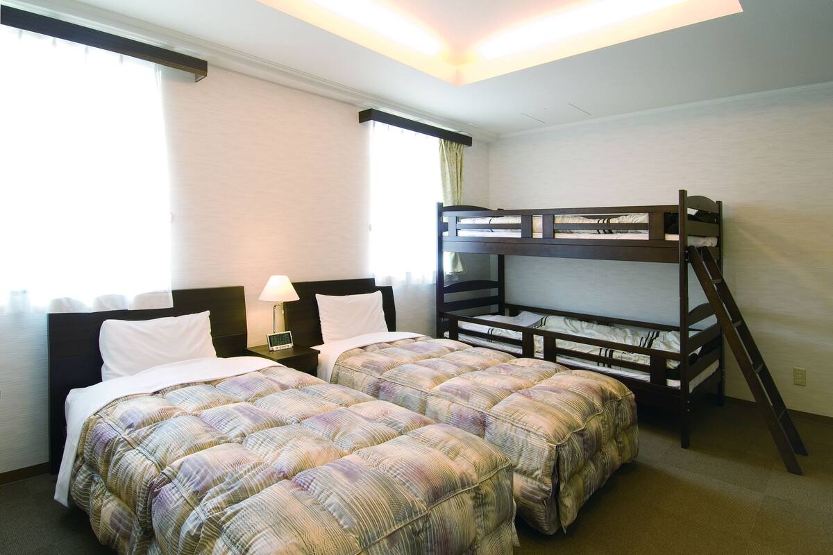 A hotel room with two single beds, a bunk bed, a nightstand with a lamp, and a window with curtains.