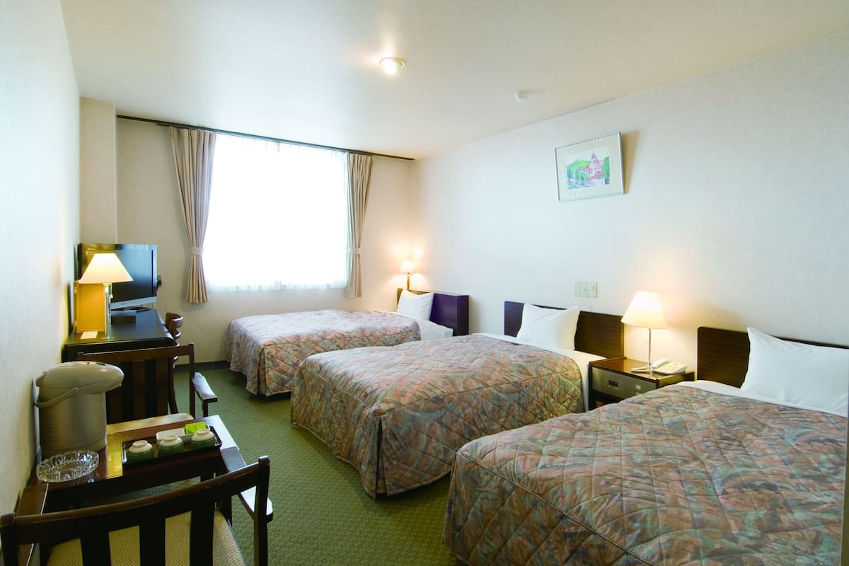 A hotel room with two single beds, a desk with a lamp, a television, and a small table with a pitcher and glasses.