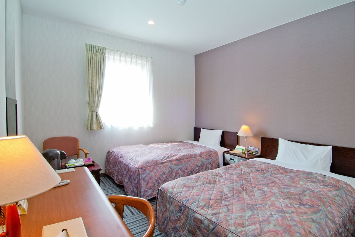 A hotel room with two beds, a desk with a lamp, a chair, a nightstand with a lamp, and a window with curtains.