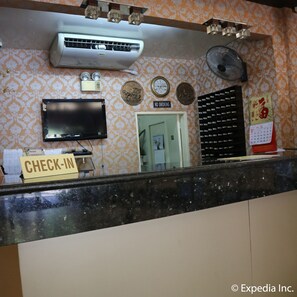 Zona de recepción de hotel con mostrador de registro, televisor, reloj de pared, calendario y un cartel de 'No Fumar'.