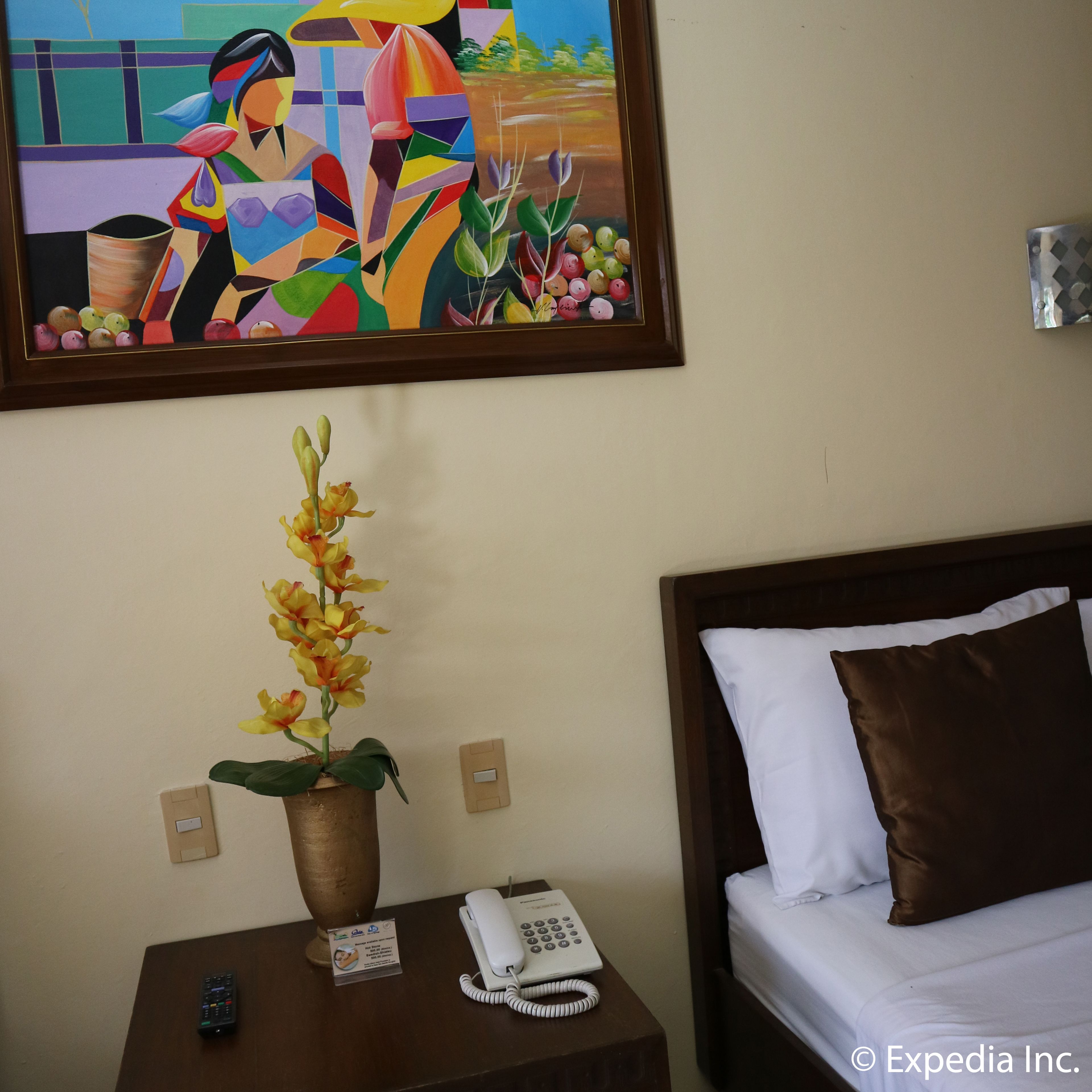 Habitación de hotel con cama, mesita de noche con teléfono, jarrón con flores y un cuadro en la pared.