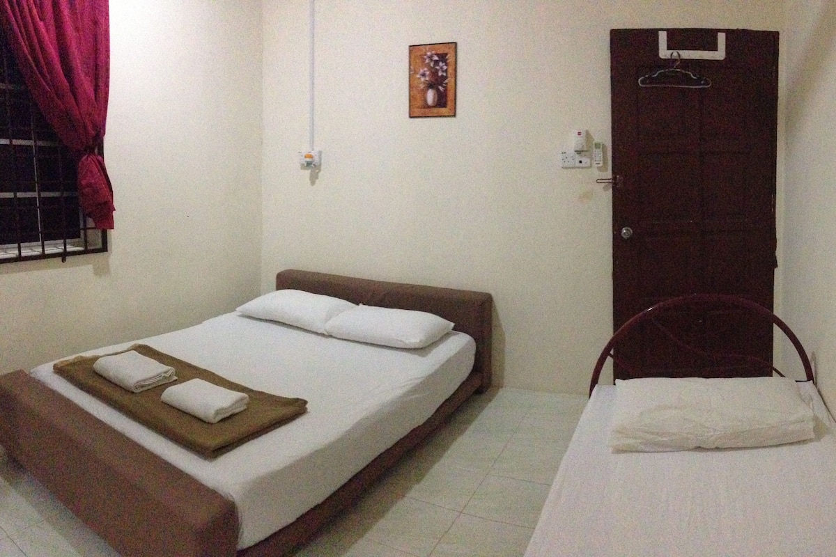 A small, single-bed room with a wooden headboard and a plain white mattress. There is a small wooden table with a folded white towel on it. The room has a simple, functional design with white walls and a window with red curtains.
