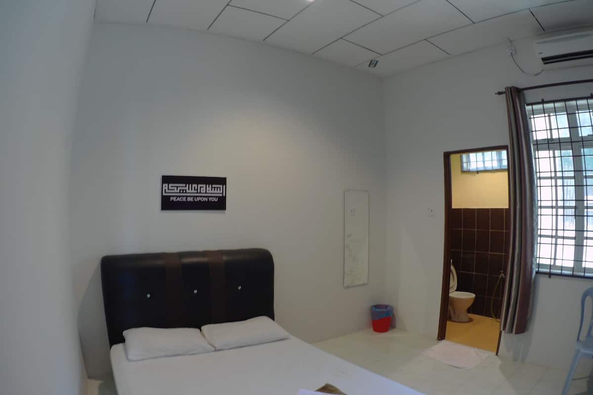 A hotel room with a bed, a wall-mounted sign, a window with curtains, a bathroom with a toilet, and a trash bin.