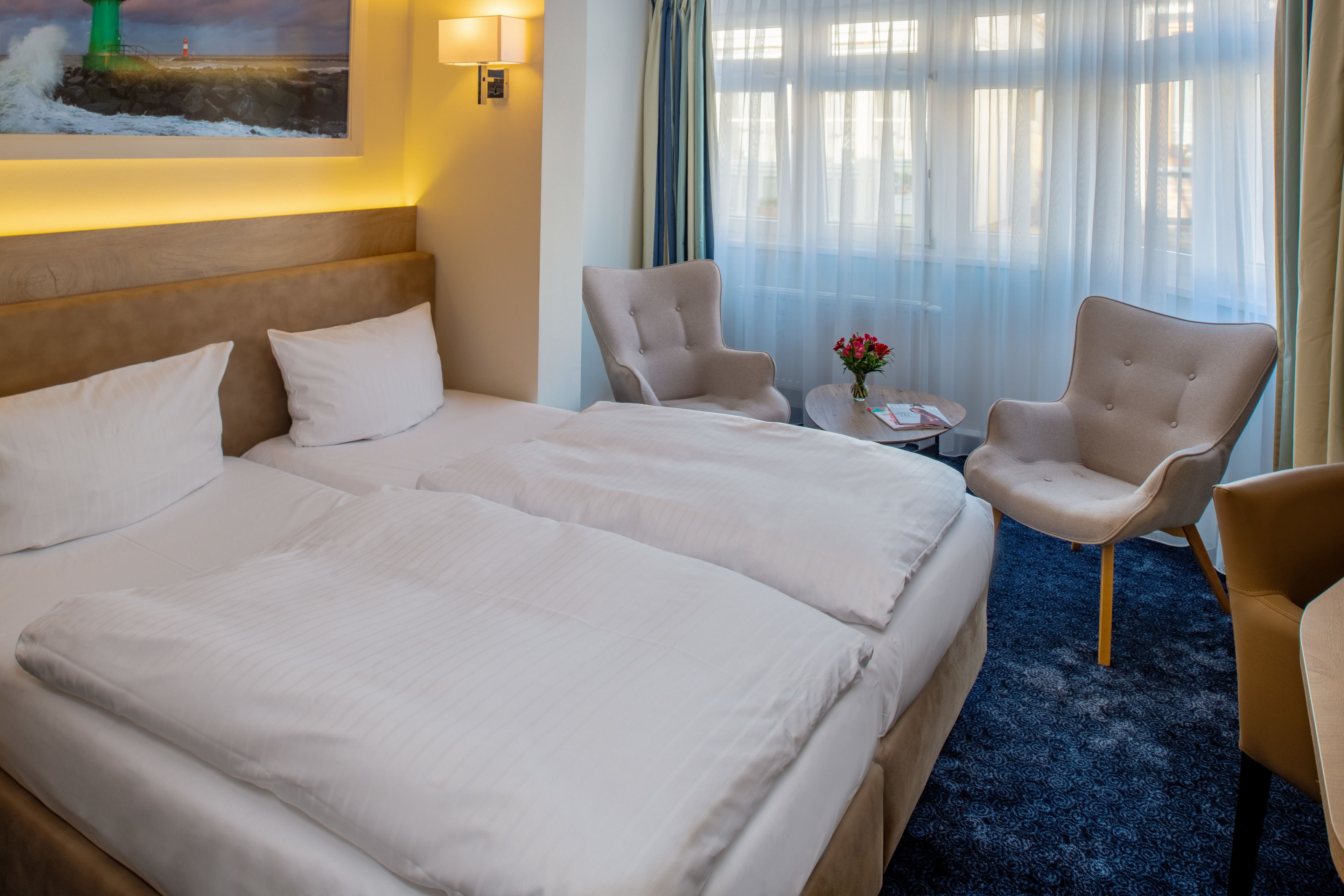 A hotel room with a large bed, two armchairs, a small table with a vase of flowers, and a painting of a lighthouse on the wall.