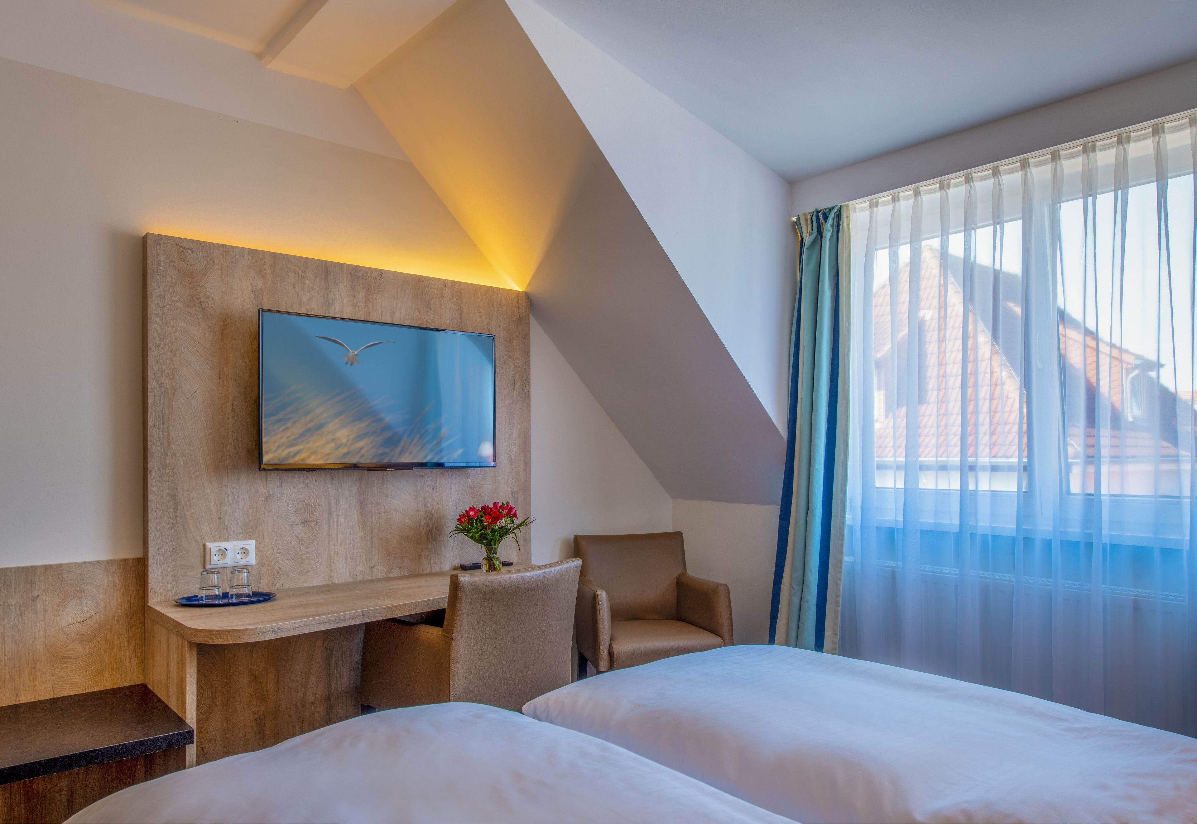 A hotel room with two beds, a desk with a chair, a TV mounted on a wooden panel, and a window with sheer curtains.