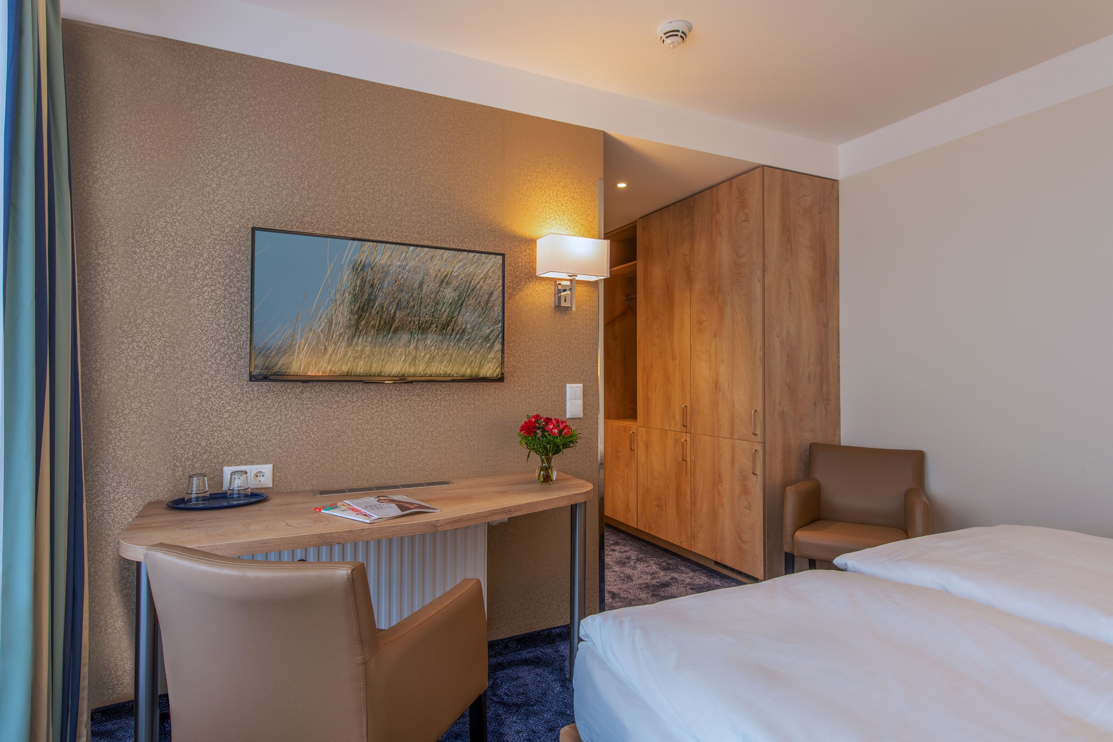 Une chambre d’hôtel comprenant un lit, un bureau avec une chaise, une armoire en bois, une lampe fixée au mur et un tableau encadré.