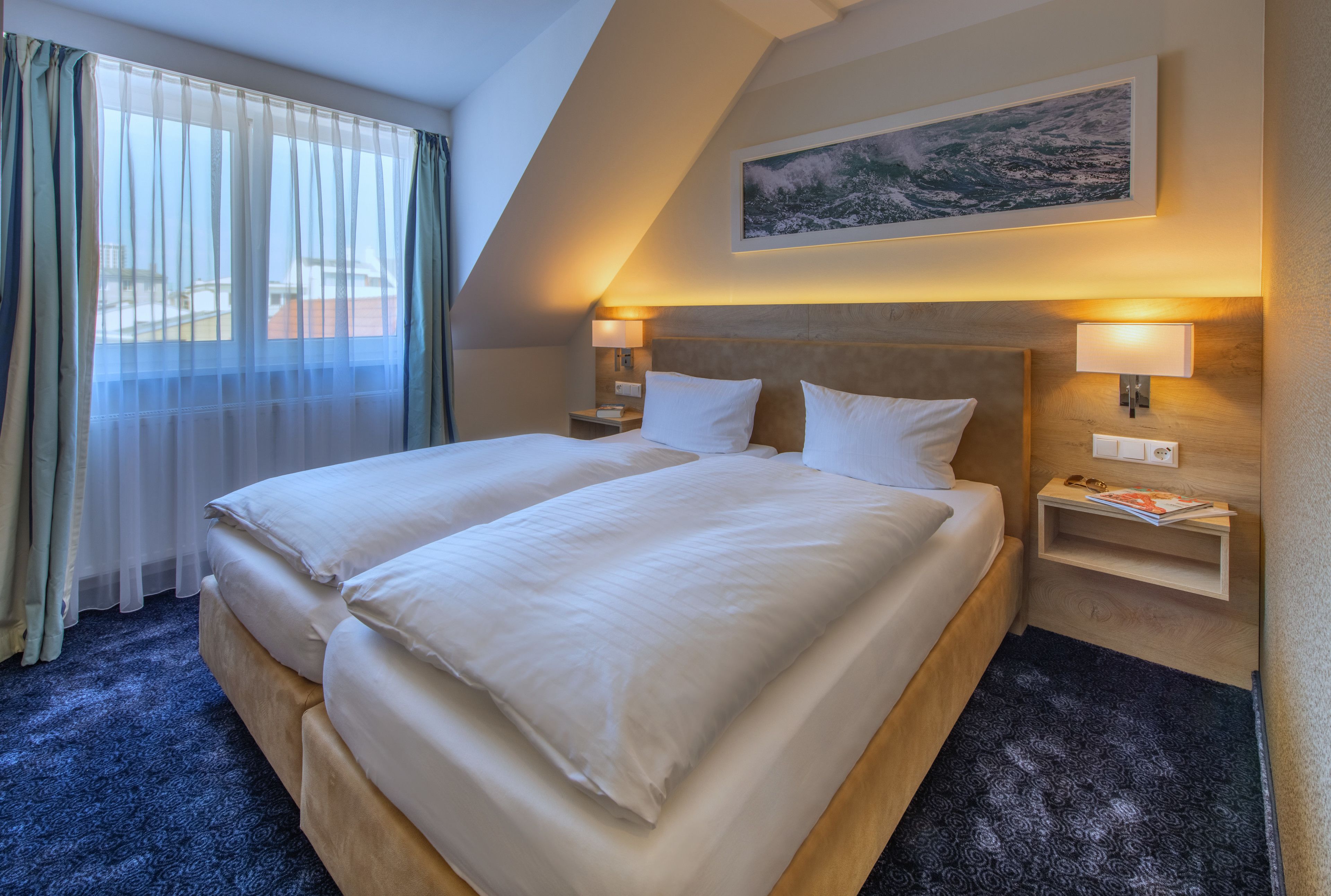 A double bed with white linens, two bedside tables with lamps, a framed picture on the wall, and a window with sheer curtains.