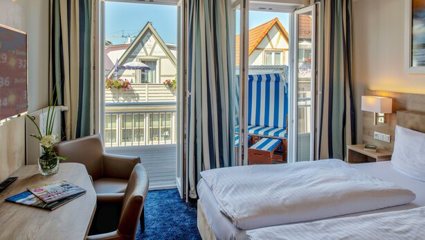 A hotel room with a bed, a desk, a chair, a small table with a vase of flowers, and a view of the outside balcony with striped curtains. - Pension StromInn (Rostock)