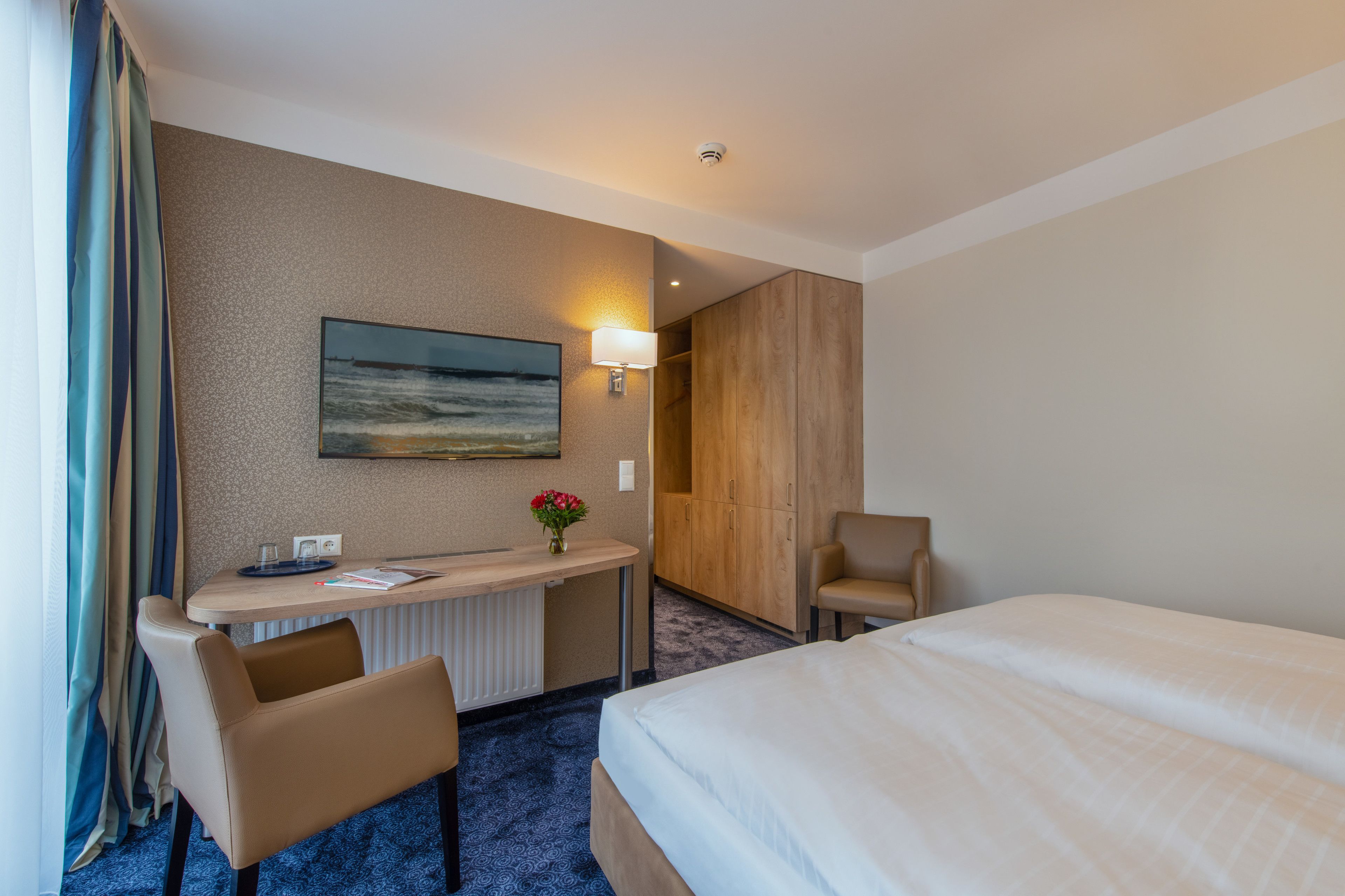 A hotel room with a bed, a desk with a chair, a lamp, a painting, and a wardrobe.