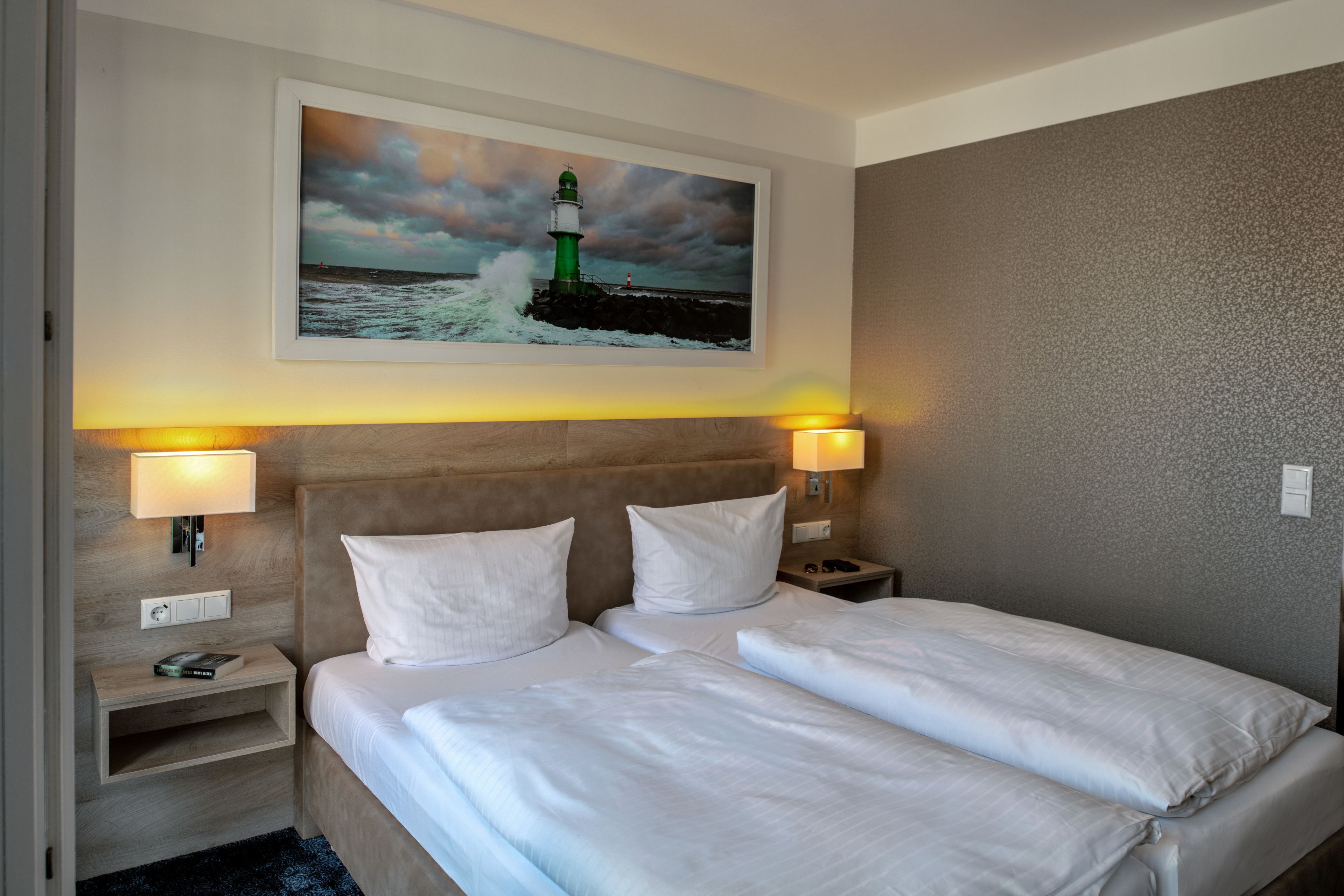 Ein Hotelzimmer mit zwei Betten, einem Nachttisch, einer Lampe und einem Gemälde eines Leuchtturms an der Wand.