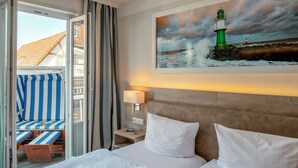 A hotel room with a bed, a nightstand, a lamp, a painting of a lighthouse, and a view of a beach chair and a building outside the window. - Pension StromInn (Rostock)