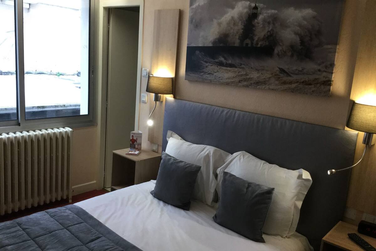 A hotel room with a large bed, two bedside lamps, a nightstand with a remote control, a radiator, and a painting of a stormy sea on the wall.