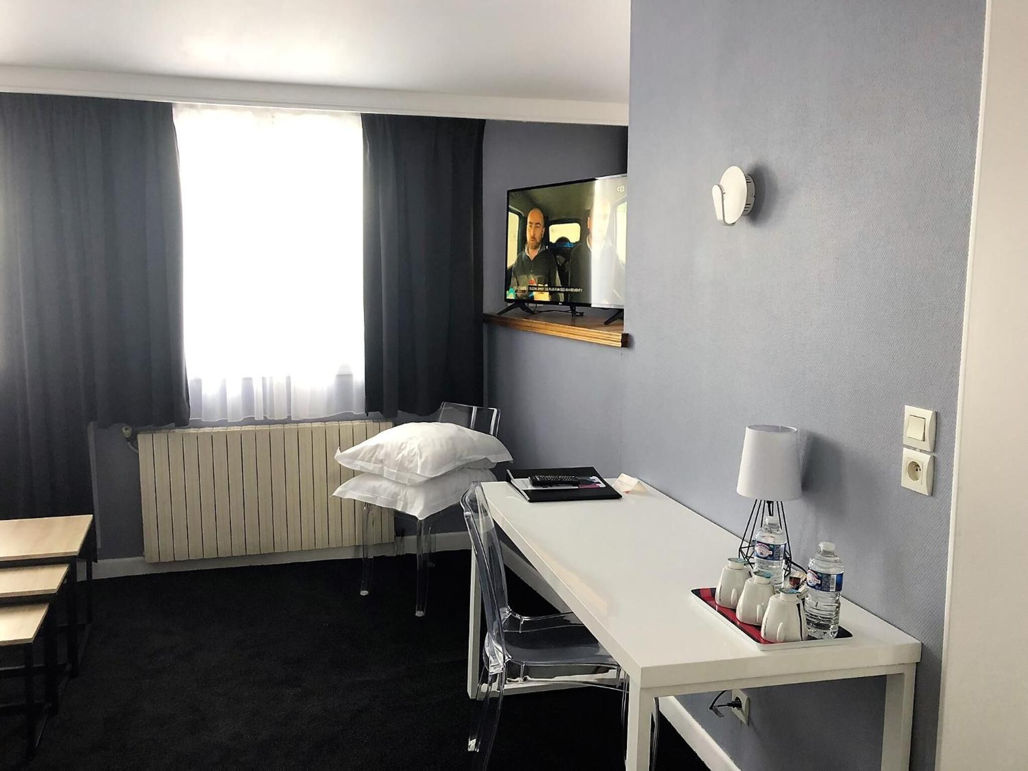 Hotelkamer met een wit bureau, een stoel, een radiator, een raam met gordijnen, een spiegel, een klok en een nachtkastje met een lamp en waterflessen.