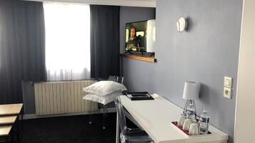 Hotelkamer met een wit bureau, een stoel, een radiator, een raam met gordijnen, een spiegel, een klok en een nachtkastje met een lamp en waterflessen.