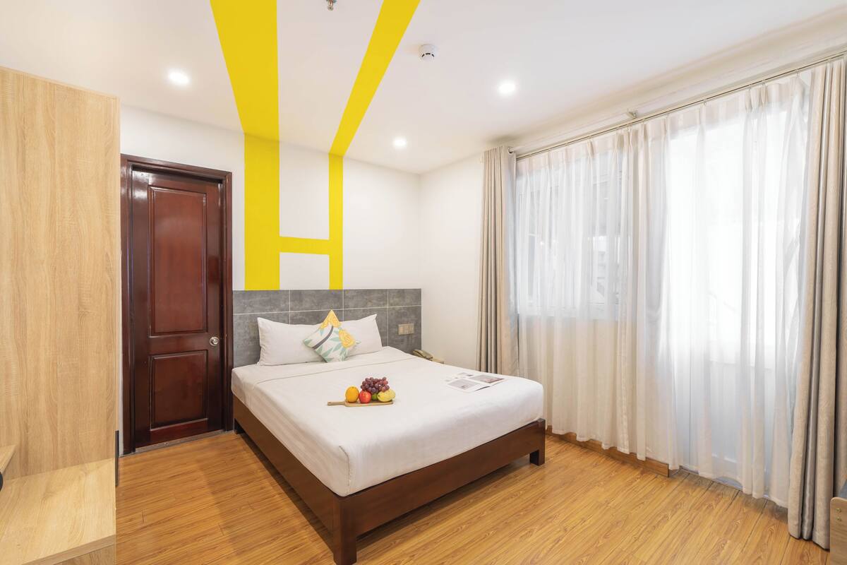 A hotel room with a wooden floor, a bed with white bedding, a wooden headboard, a door, and a window with curtains.