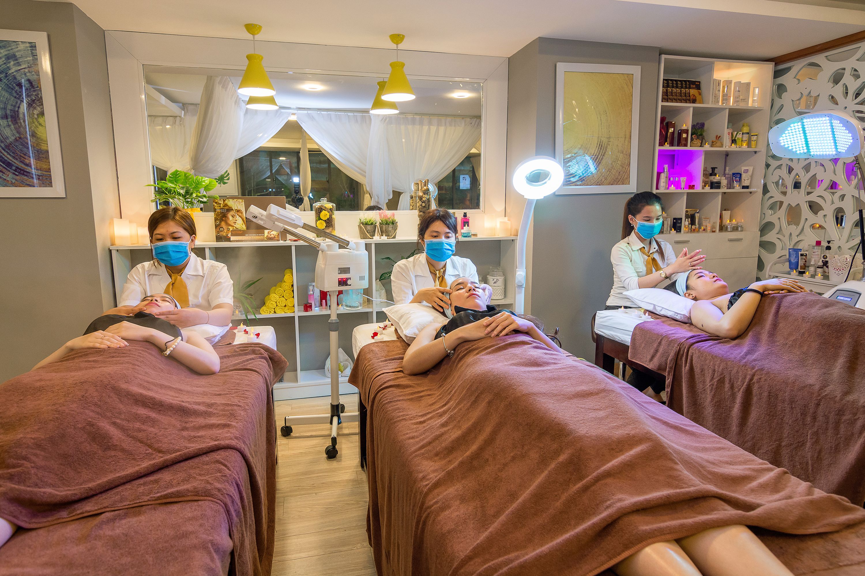 A spa treatment room with three clients receiving massages, a well-lit reception area, and a shelf with various products.