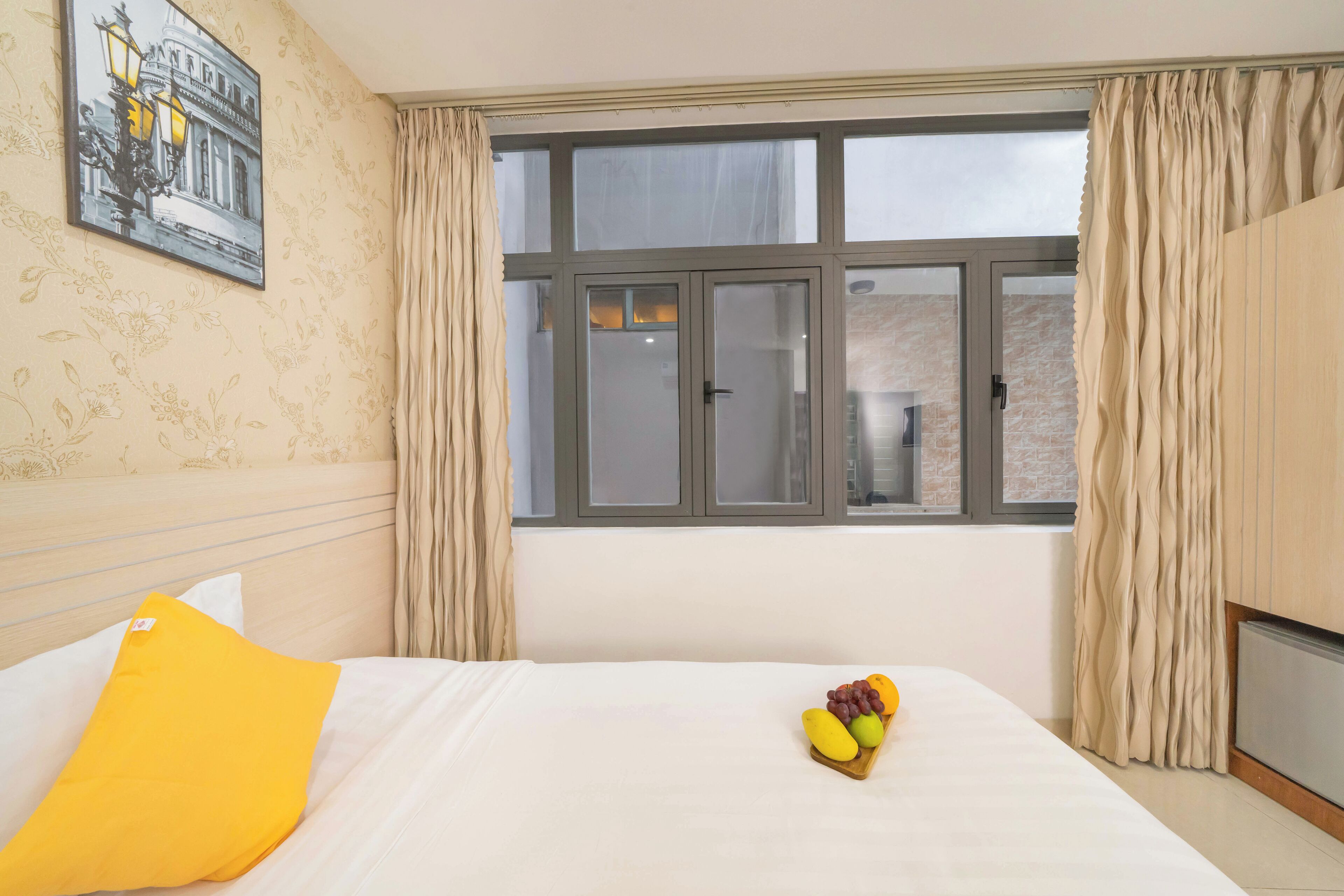 A hotel room with a bed, a window with curtains, a wall-mounted picture, and a fruit basket on the bed.