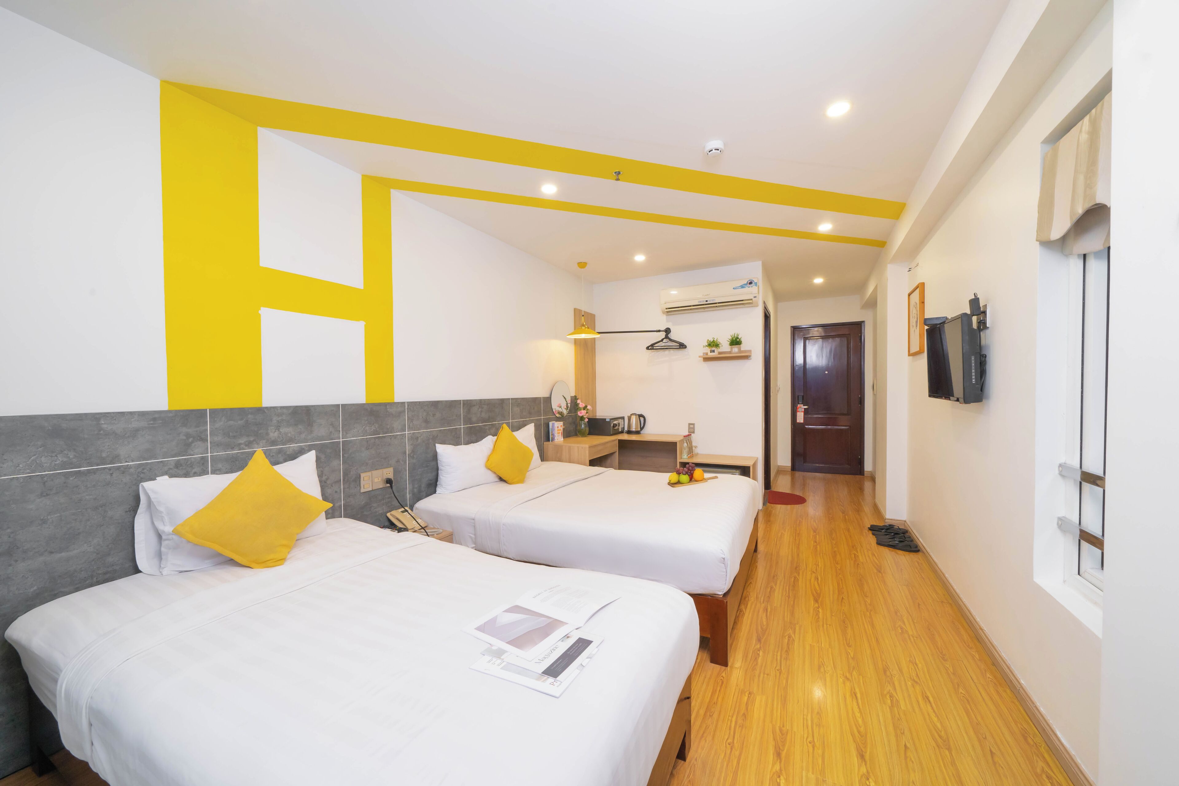 A hotel room with two beds, a wooden floor, a small kitchenette, and a TV on the wall.