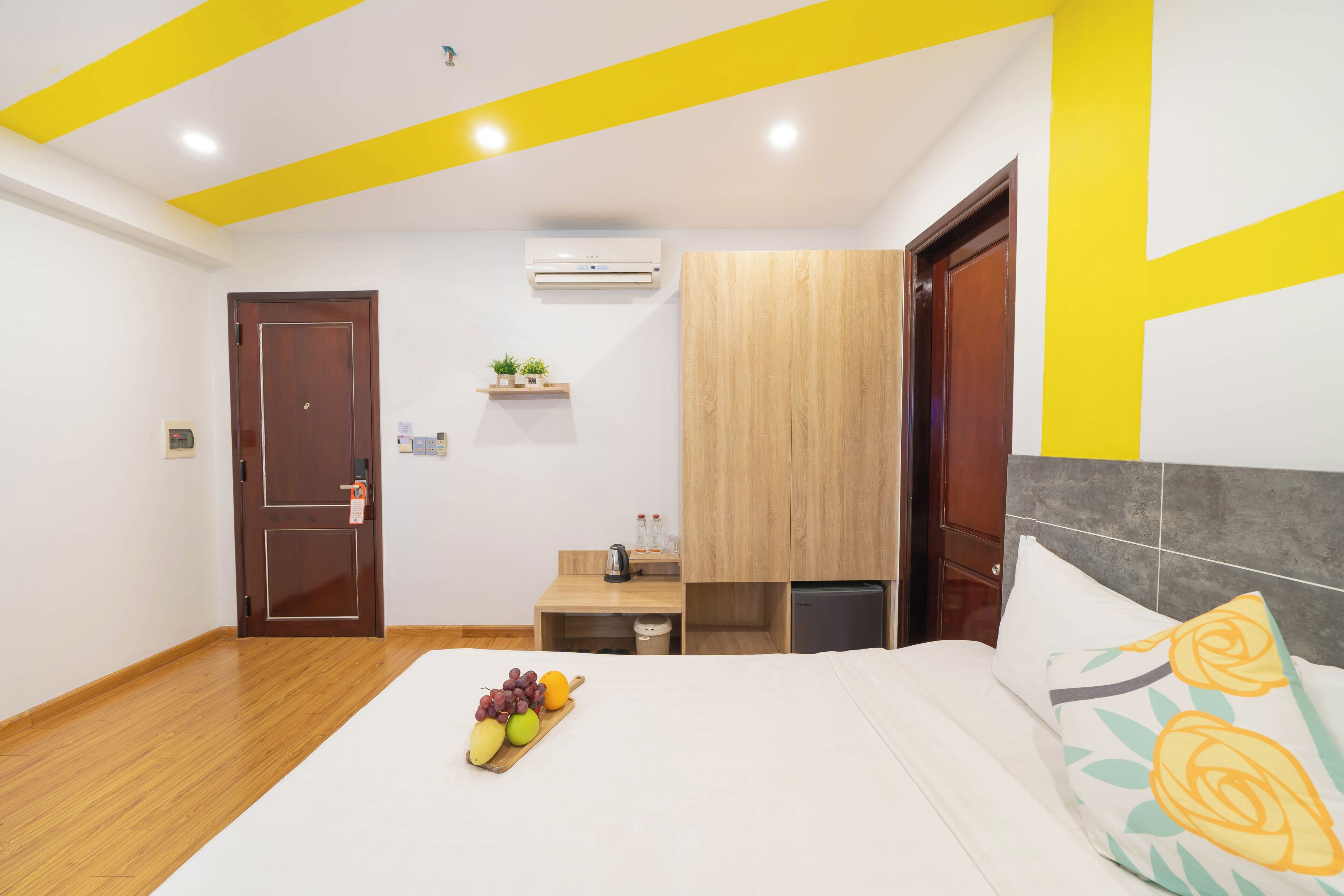 A hotel room with a wooden floor, a bed with a white bedspread, a bedside table with a fruit bowl, a small wooden nightstand, a wall-mounted air conditioner, and a wooden wardrobe.