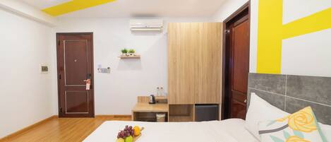 A hotel room with a wooden floor, a bed with a white bedspread, a bedside table with a fruit bowl, a small wooden nightstand, a wall-mounted air conditioner, and a wooden wardrobe.