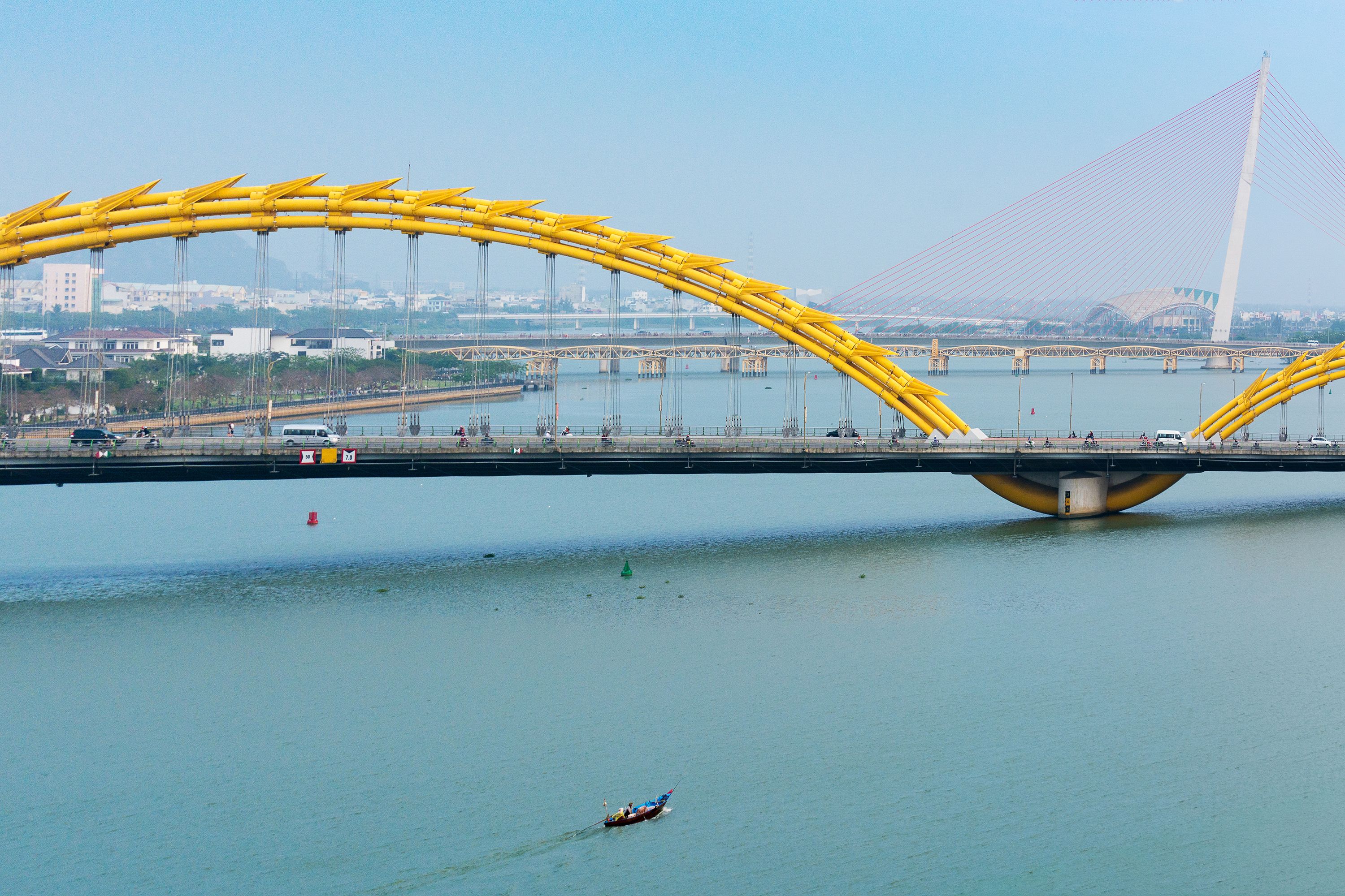 Un puente con un diseño distintivo de arco amarillo que cruza un cuerpo de agua, con otro puente atirantado al fondo.