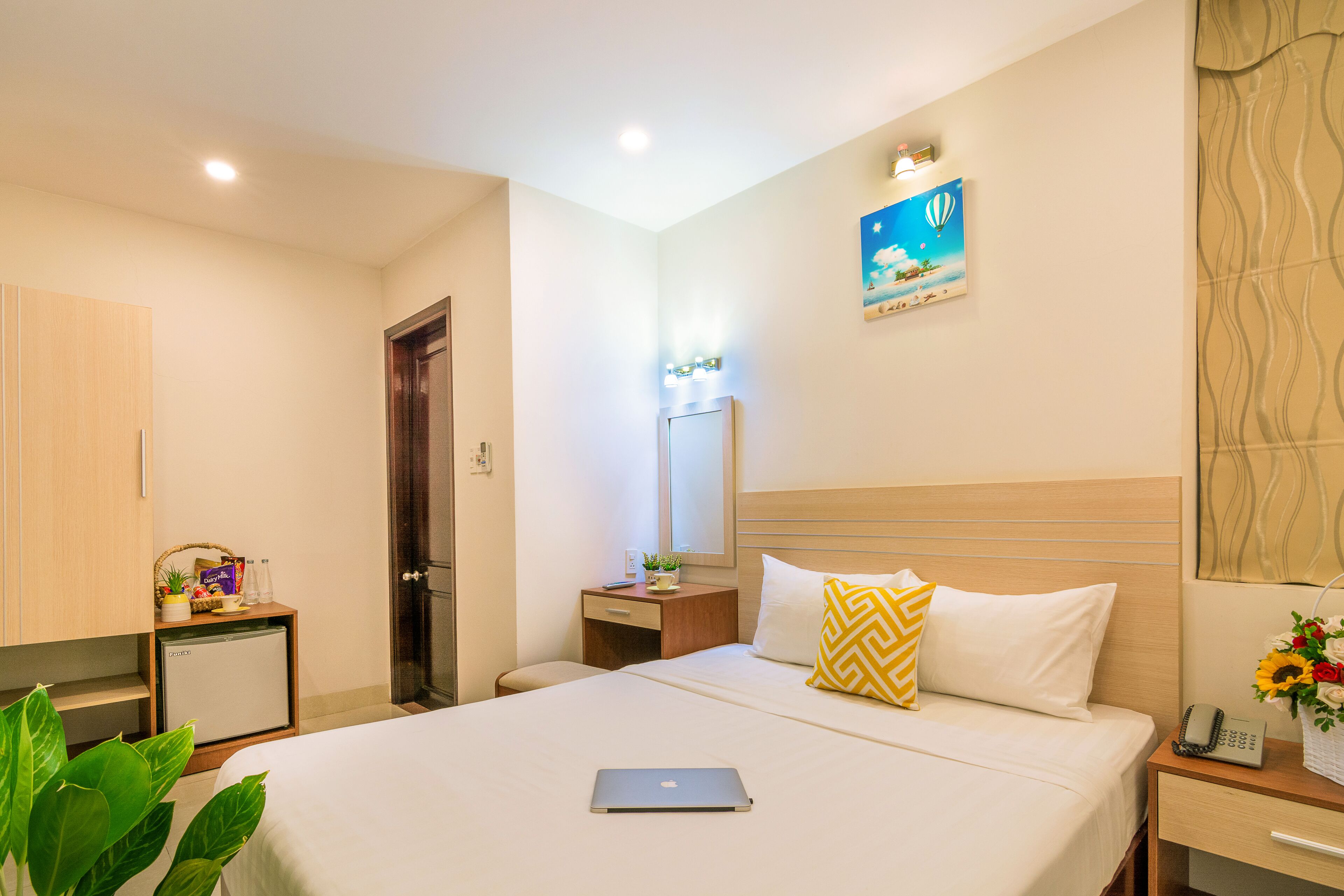 A hotel room with a bed, a desk with a laptop, a small refrigerator, a nightstand with a phone, and a wall-mounted mirror.
