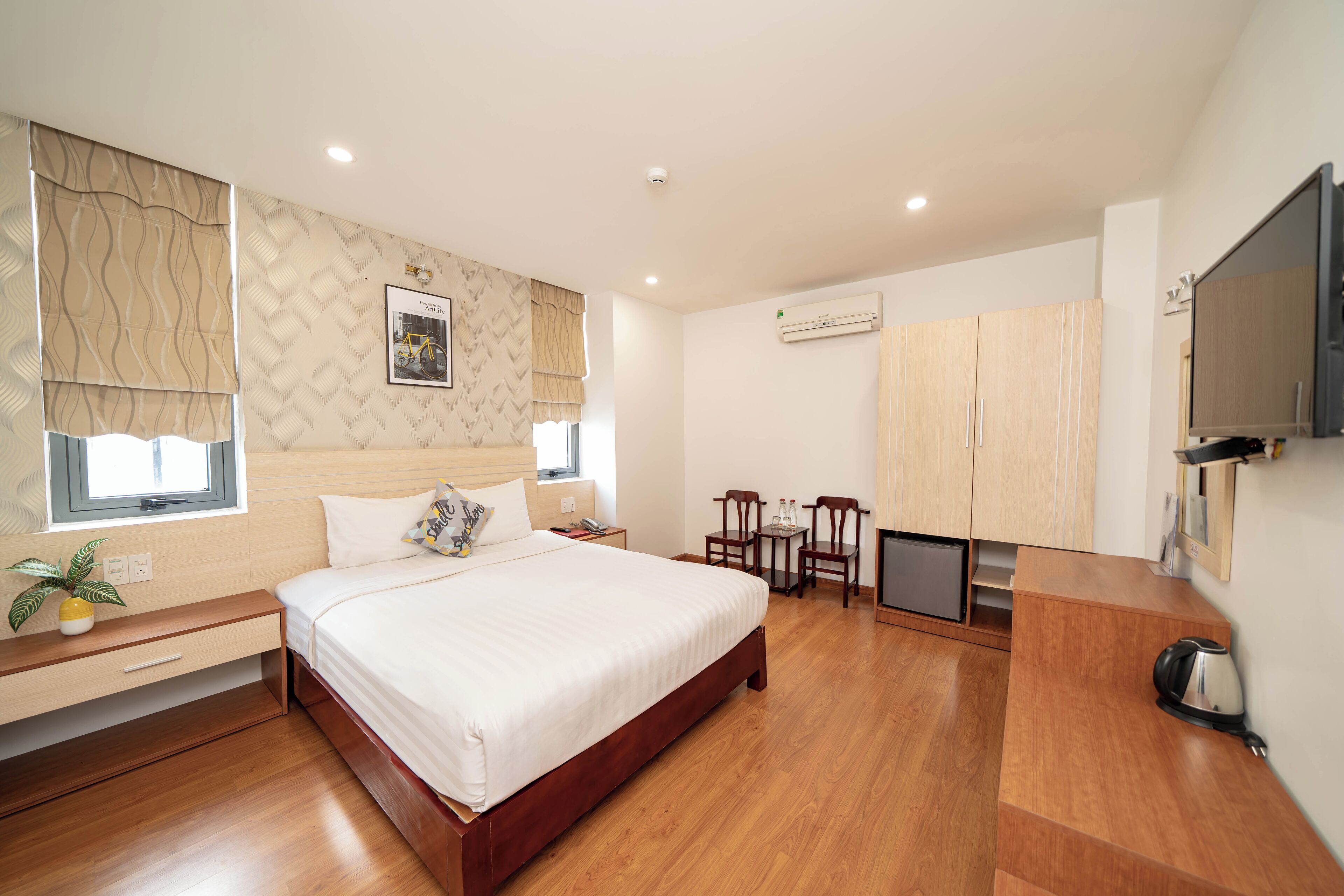 A modern hotel room with a large bed, wooden flooring, a TV, a desk with a chair, and a small kitchenette.