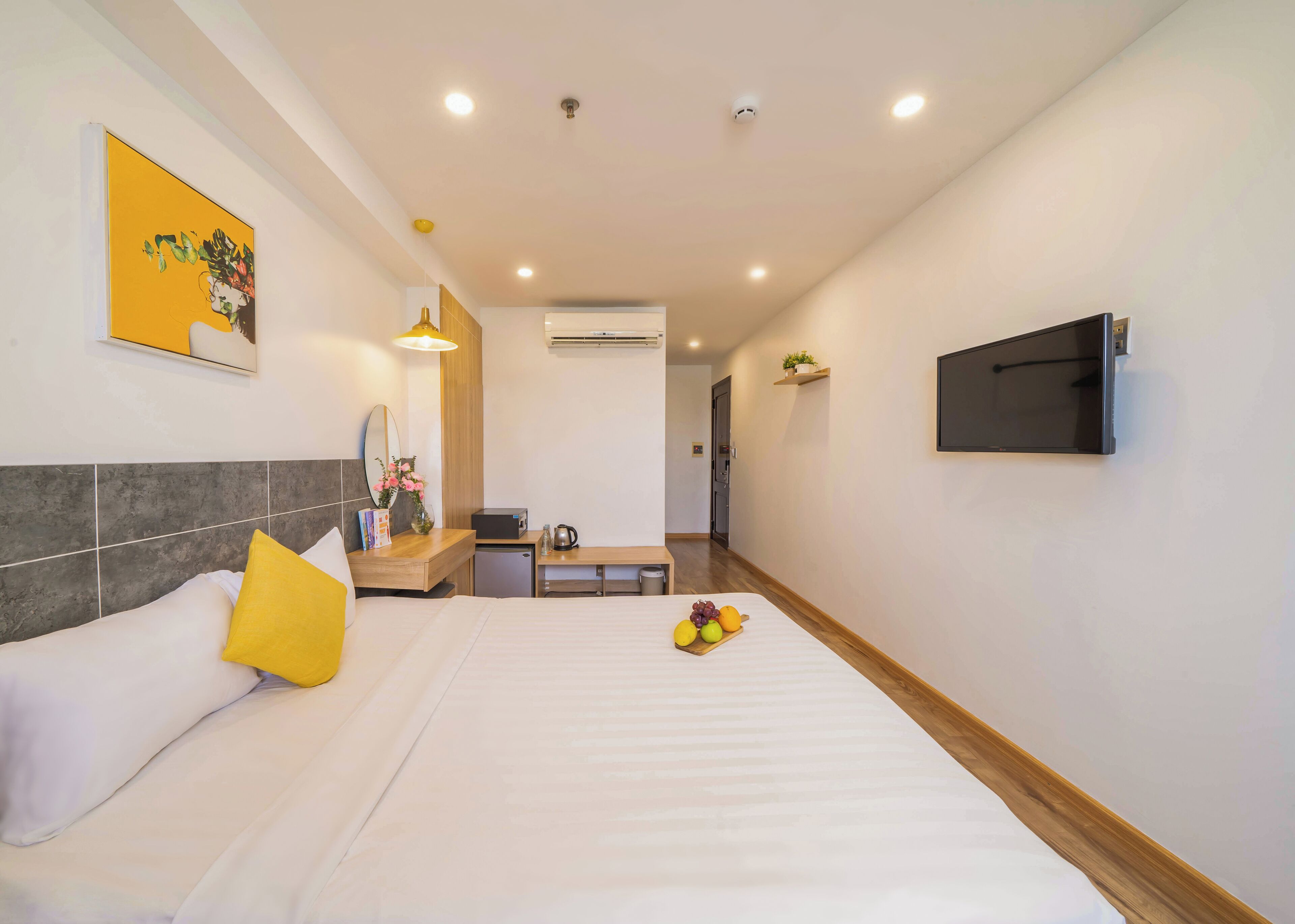 A hotel room with a bed, bedside table, a small desk, a chair, a TV mounted on the wall, and a hallway leading to another room.