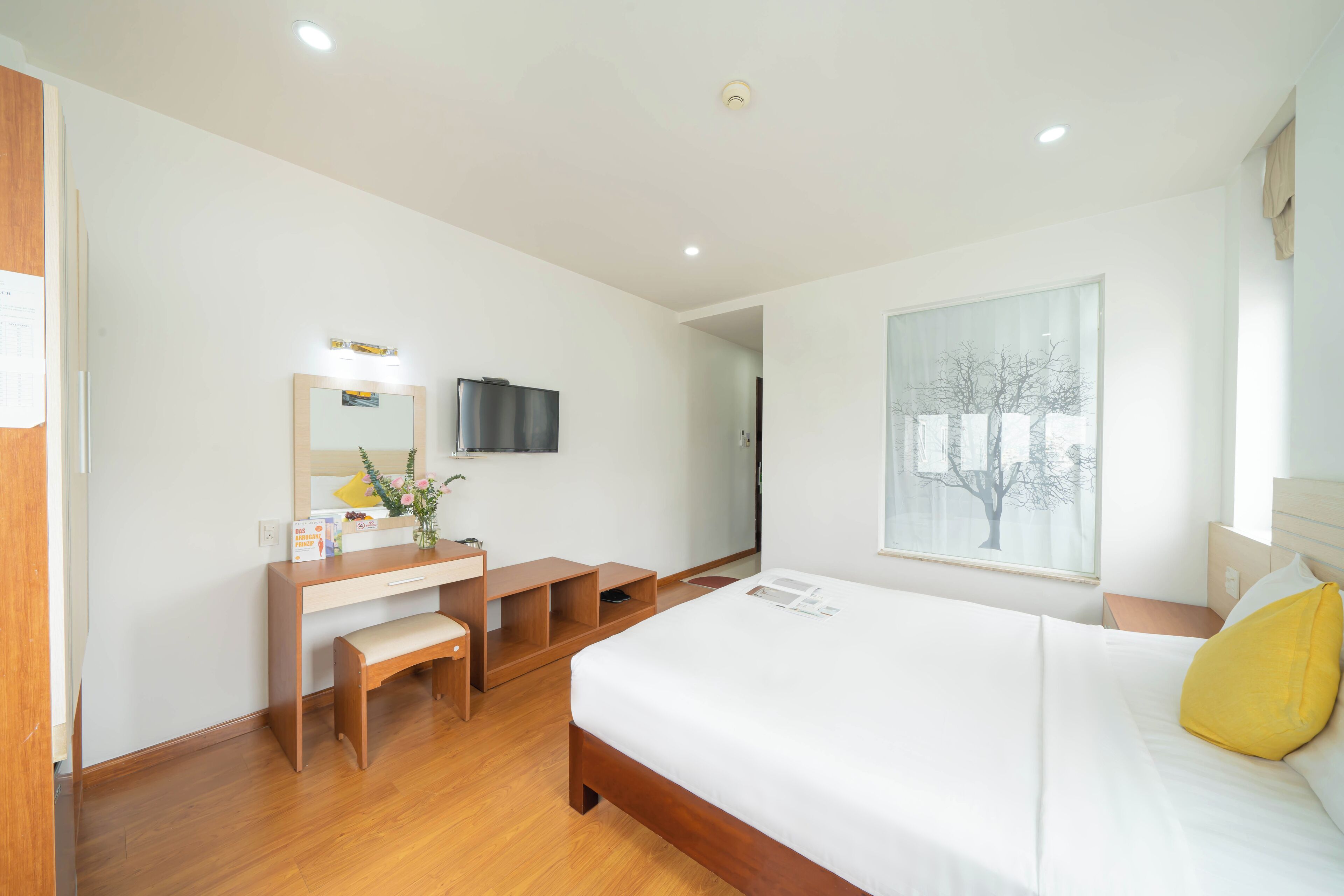 A modern hotel room with a wooden desk, a bed with white bedding, a large window with a view of trees, and a mounted flat-screen TV.