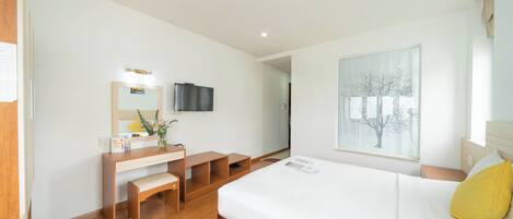 A modern hotel room with a wooden desk, a bed with white bedding, a large window with a view of trees, and a mounted flat-screen TV.