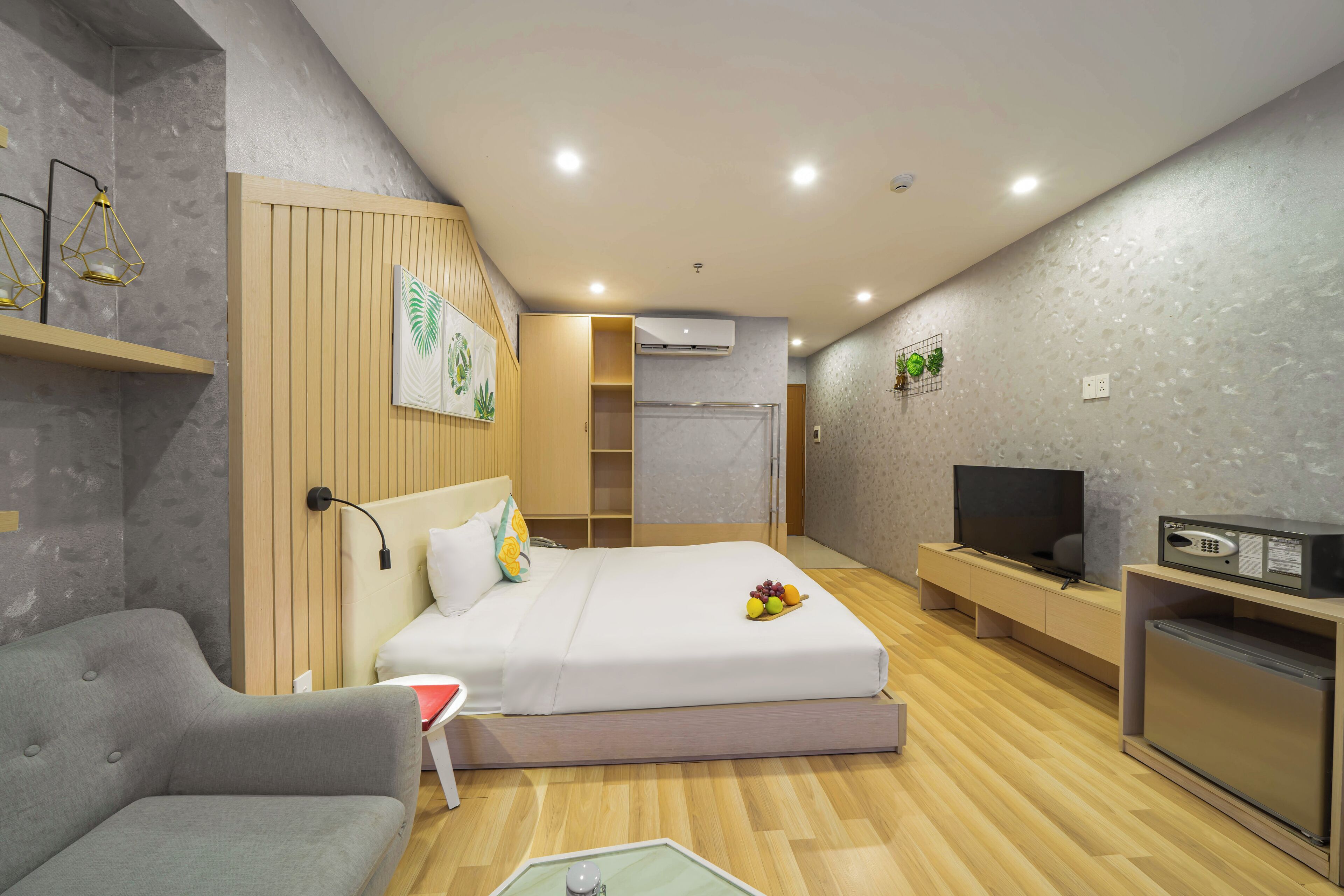 A modern hotel room with a bed, a sofa, a TV, a mini-fridge, and a desk with a lamp.