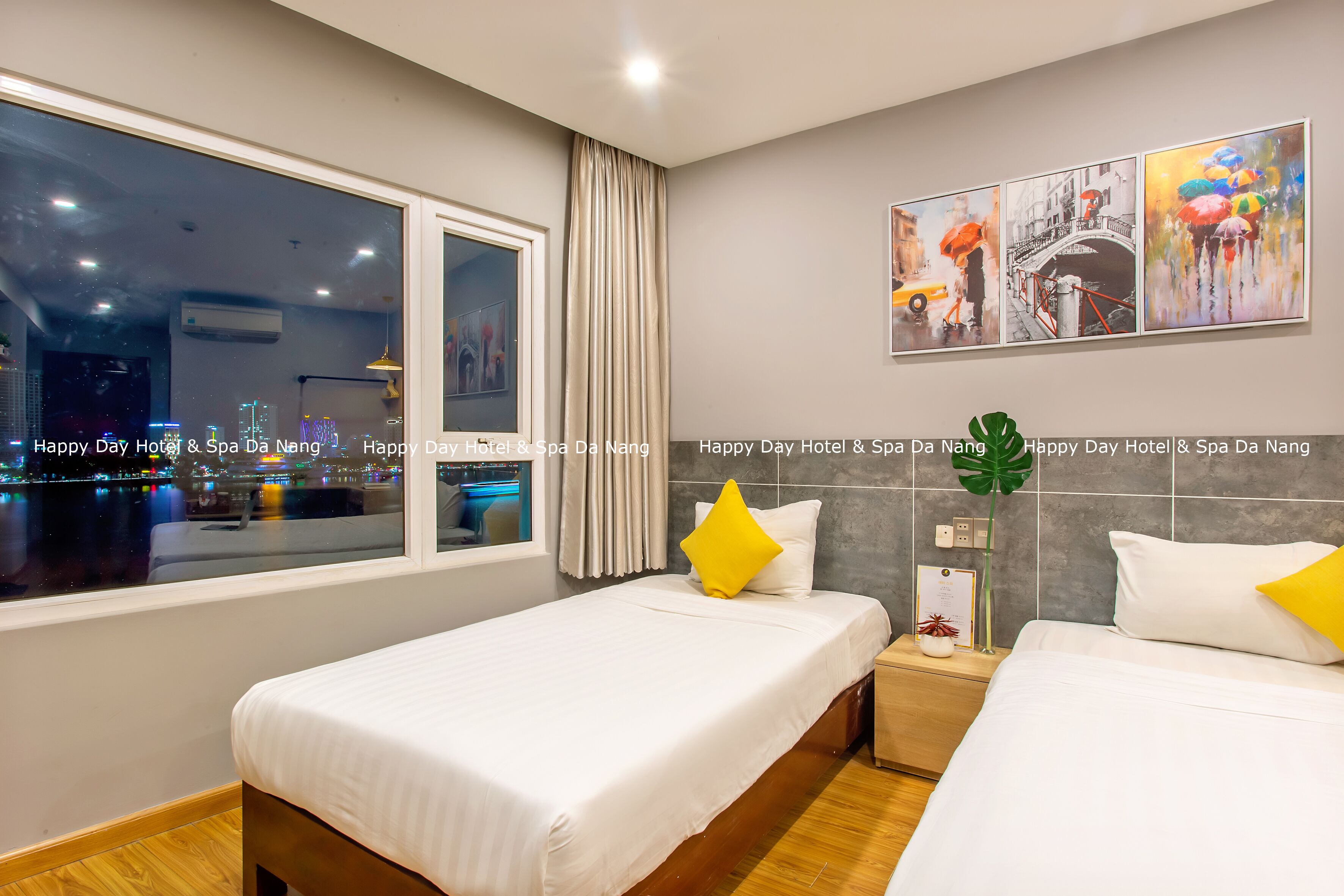 A hotel room with two beds, a large window with a city view, and artwork on the wall.