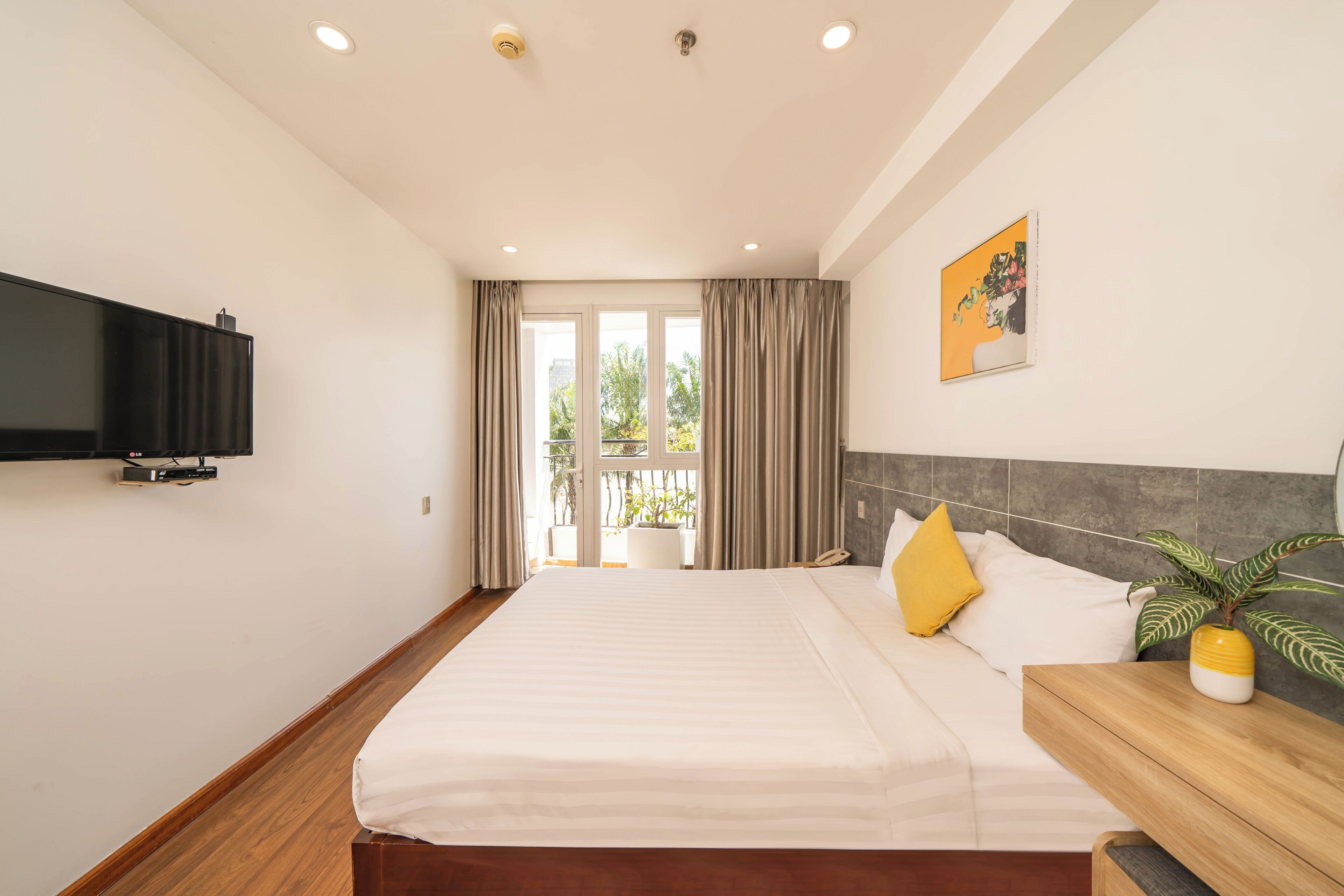 A hotel room with a large bed, a flat-screen TV mounted on the wall, a wooden bedside table with a plant, and a balcony with curtains.