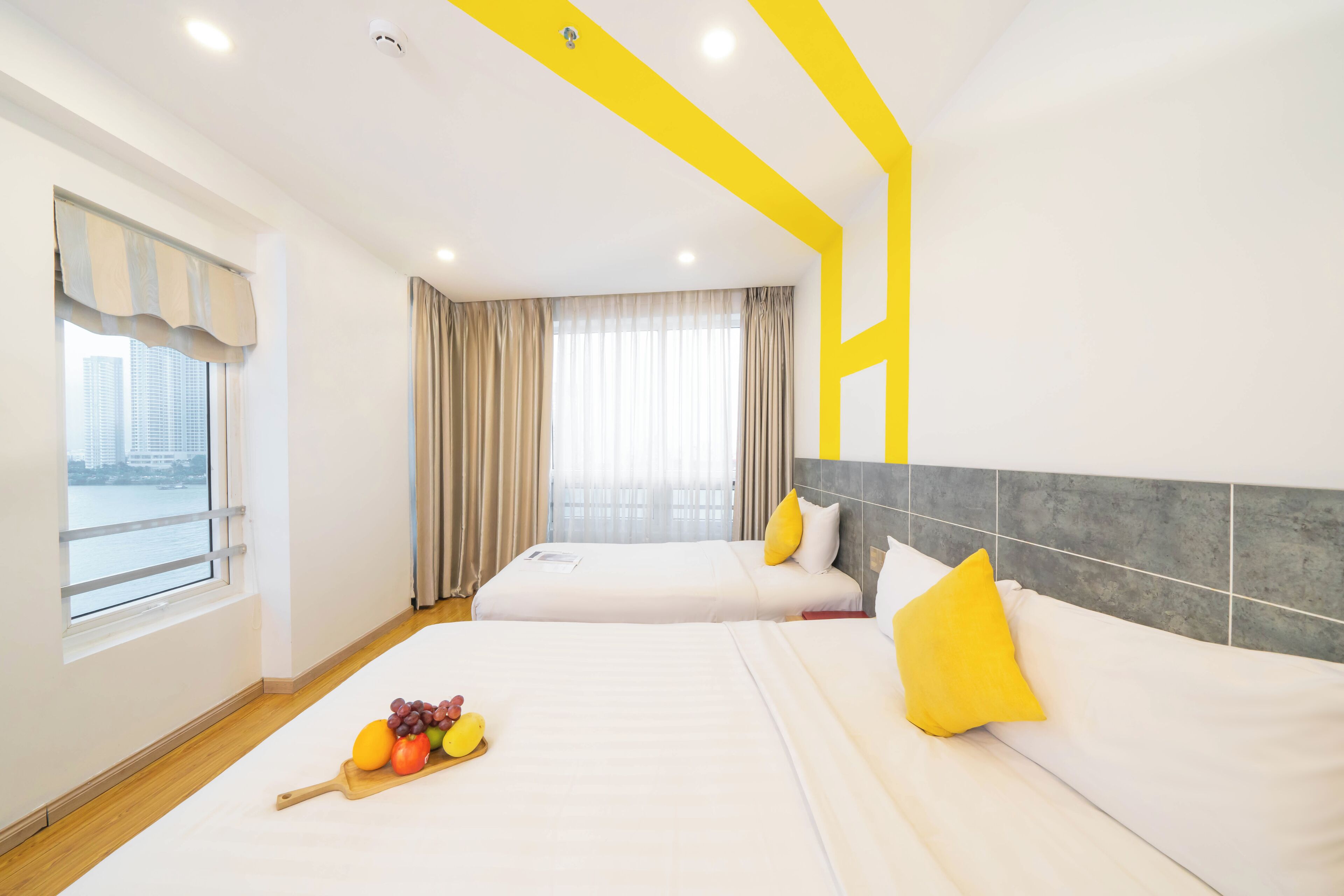 Habitación de hotel con dos camas, un ventanal con cortinas, una bandeja de frutas en el suelo y vistas a la ciudad.