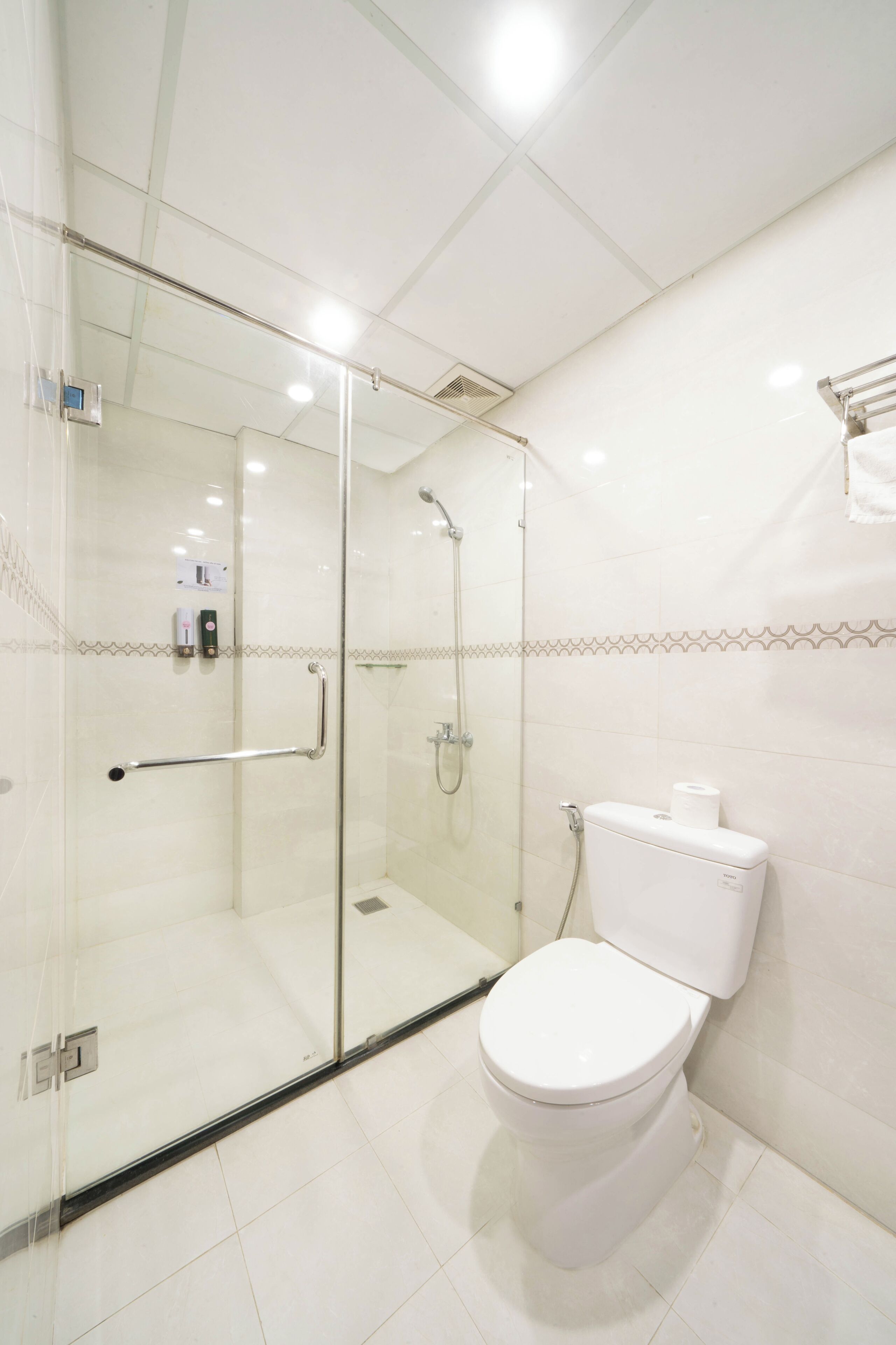 Un baño moderno con ducha envuelta en vidrio, inodoro blanco y dispensador de gel antibacterial en la pared.