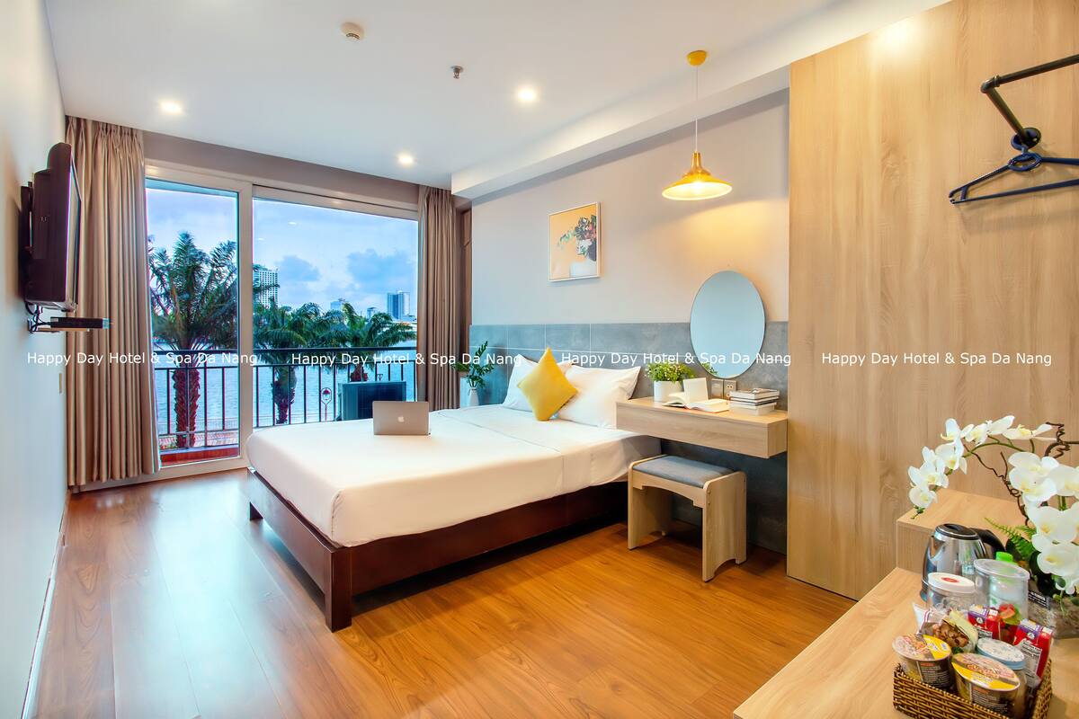 A hotel room with a large bed, a desk, a chair, a TV, and a balcony with a view of palm trees and buildings.