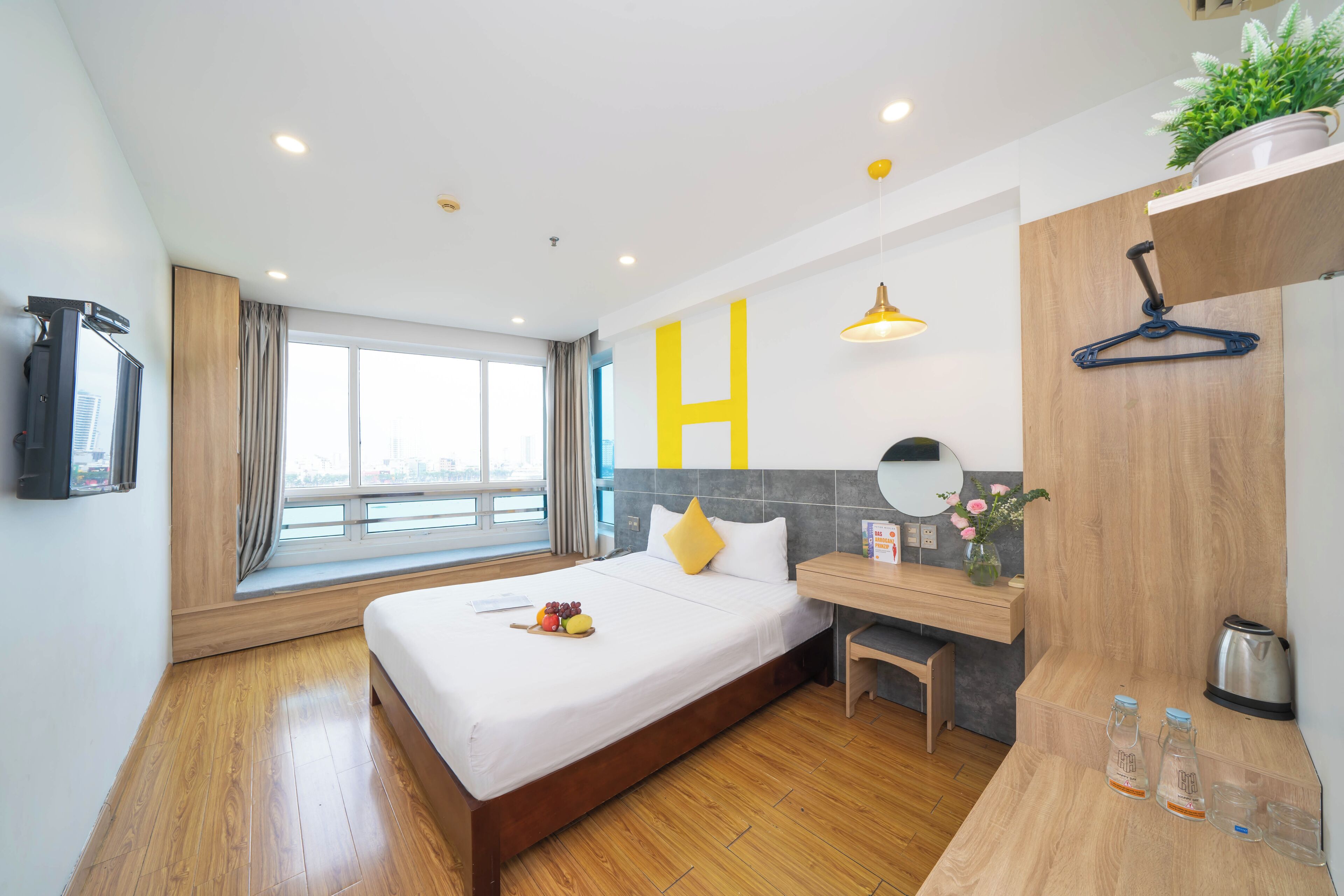 A modern hotel room with a large bed, wooden flooring, a desk with a chair, a small table with a vase of flowers, a hanging plant, and a large window with curtains.