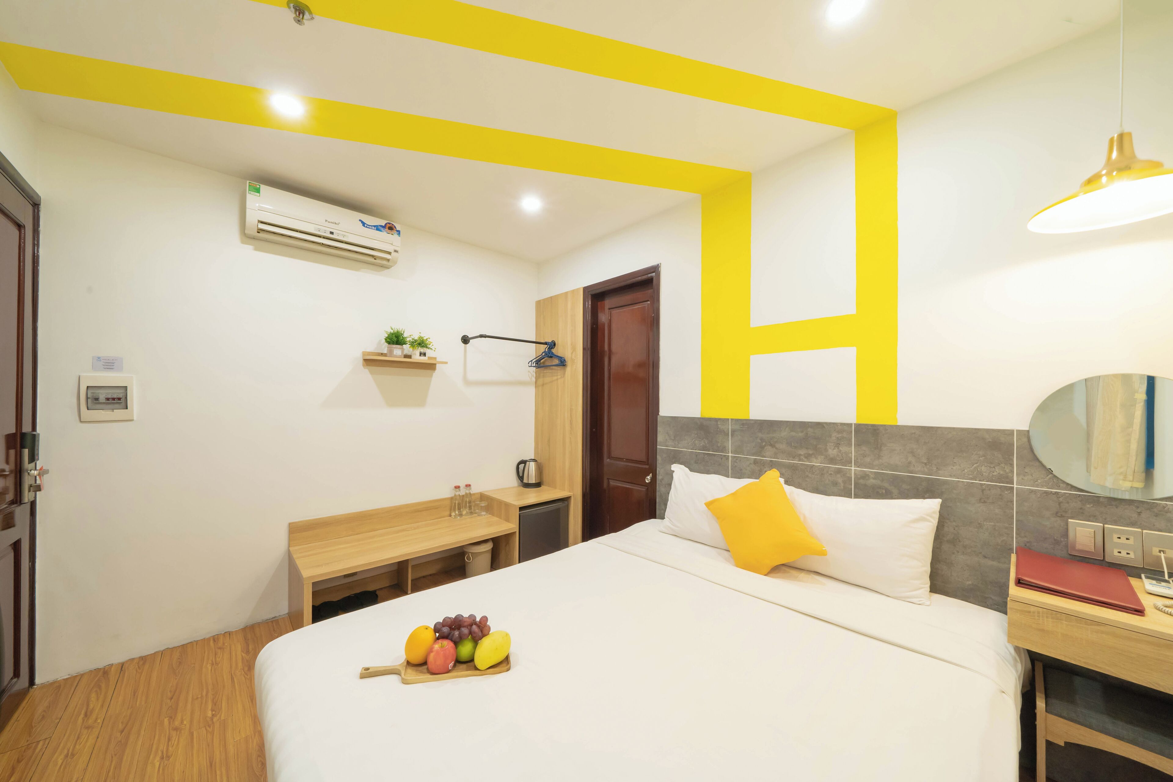 A hotel room with a bed, bedside table, and a small desk with a chair. There is a fruit tray on the bed, a mirror on the wall, and an air conditioning unit above the desk.