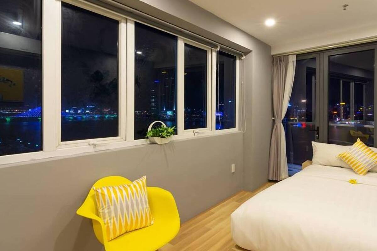 A hotel room with a bed, a chair, and large windows offering a city view at night.