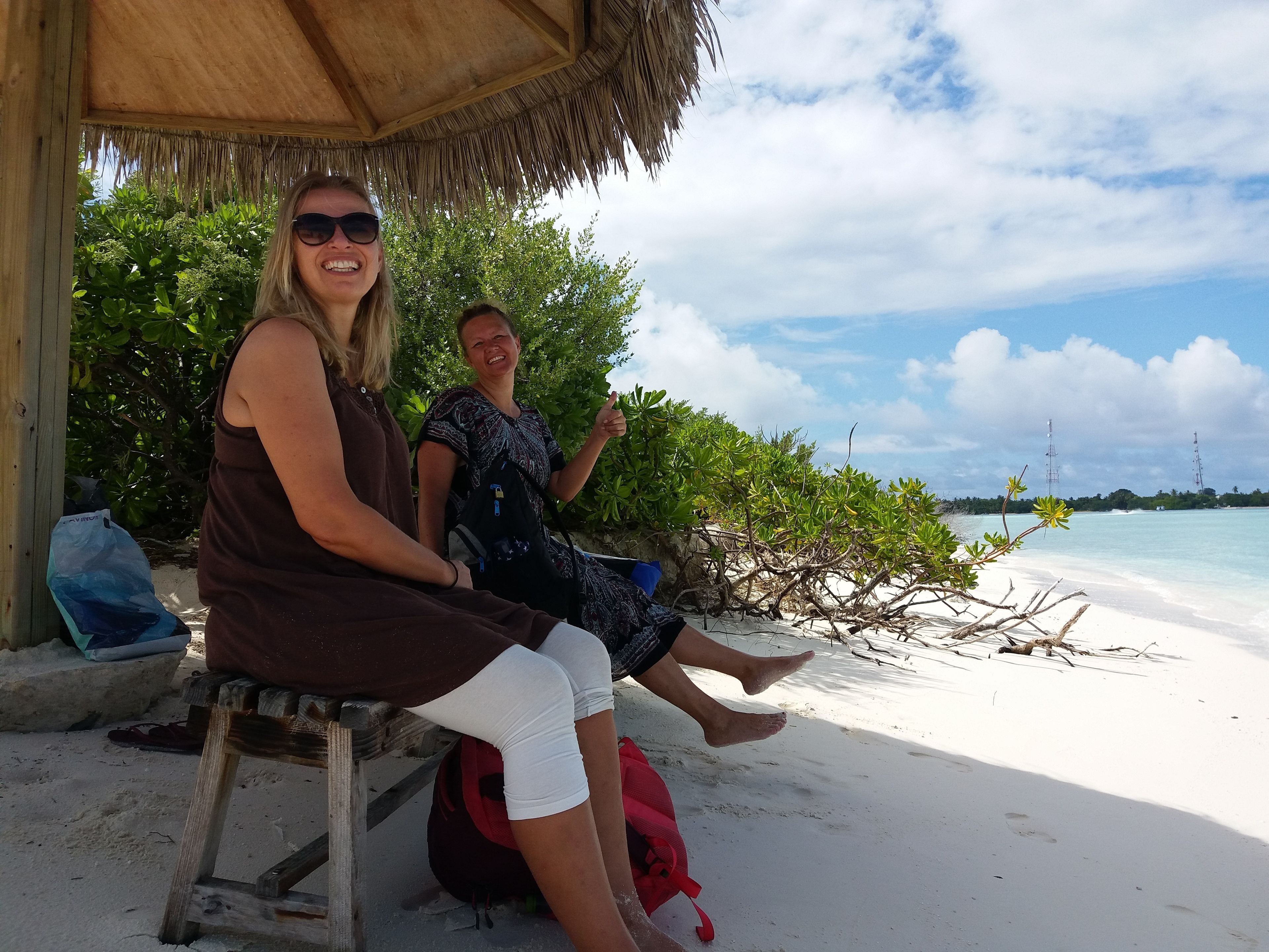 Two people sitting on a wooden bench under a thatched roof, with a beach and clear sky in the background.