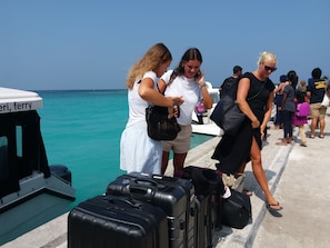 A pier with people and luggage, a boat with 'ferry' written on it, and a clear blue sea in the background.
