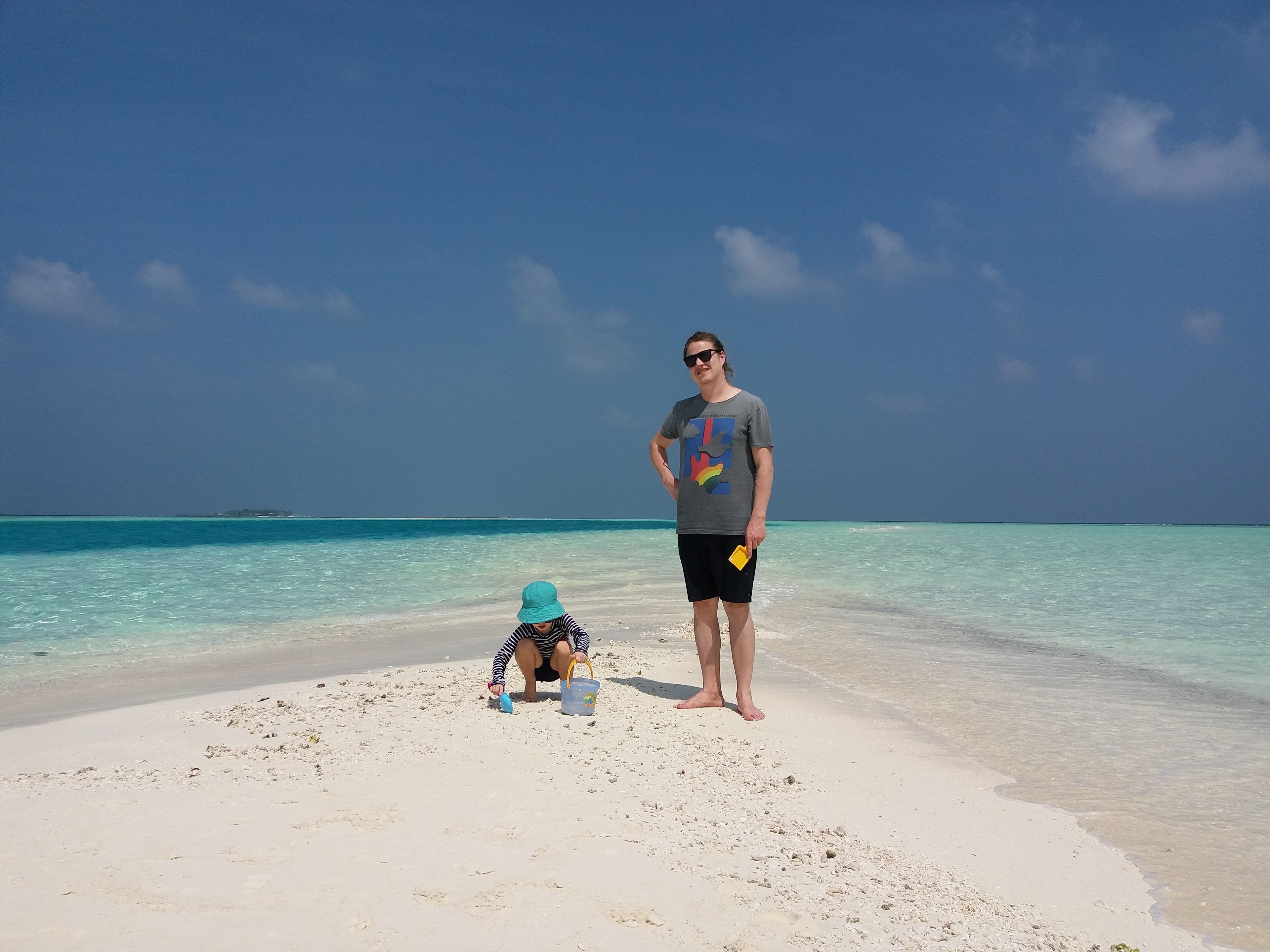 A beach scene with a person standing and a child playing with a bucket and shovel.