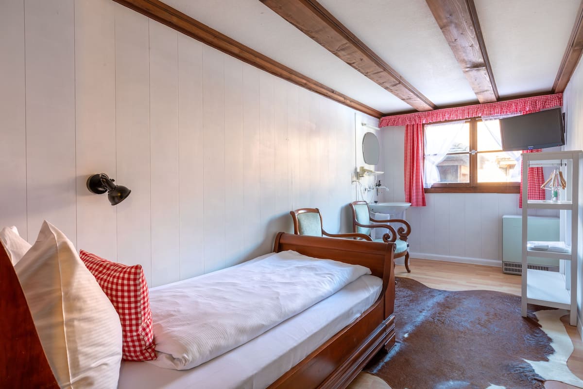 Un lit simple avec un cadre en bois, une petite table de chevet, une chaise, un bureau et une fenêtre avec un rideau à carreaux rouges.
