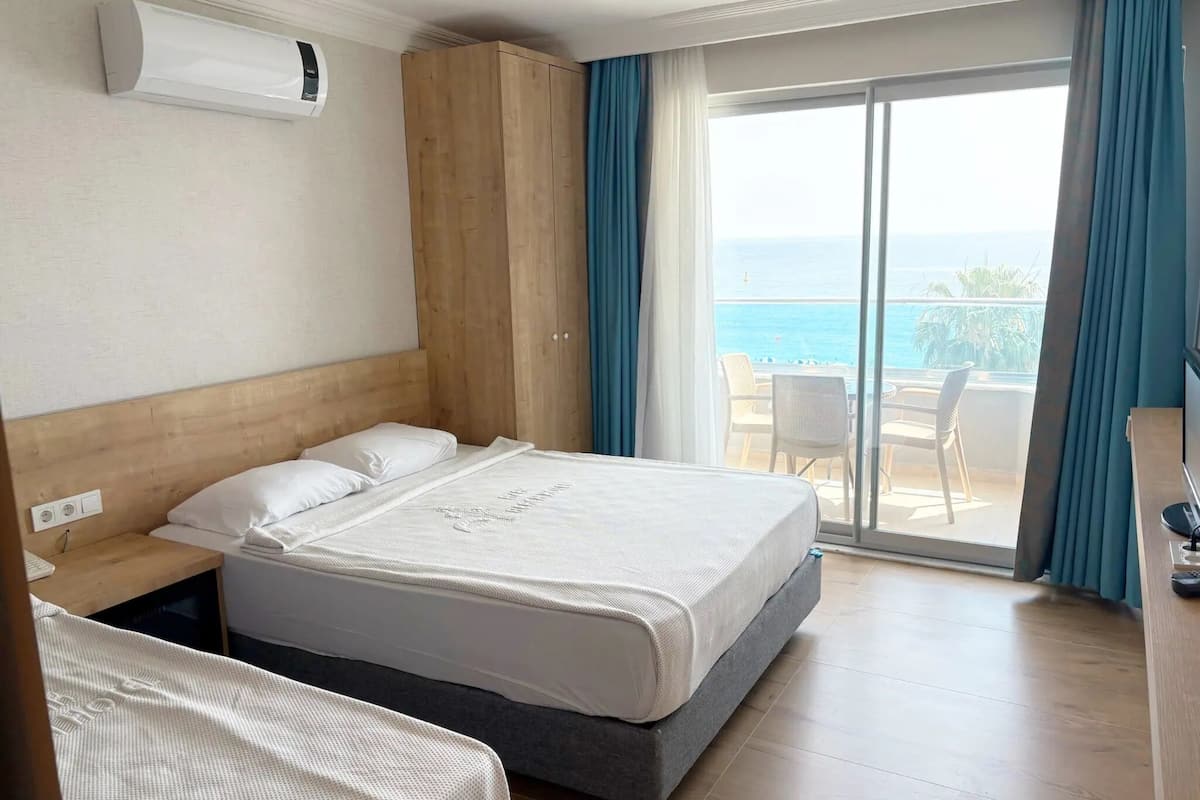 A hotel room with a bed, a wooden headboard, a window with a view of the sea, and a balcony with chairs.