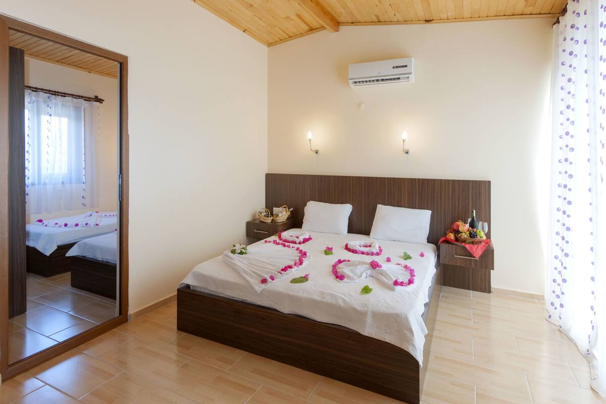 Una cama bien tendida con una colcha blanca, un cabecero de madera y una mesita de noche con una cesta de frutas.
