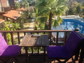 A wooden deck with two purple chairs and a table, overlooking a swimming pool and lush greenery.